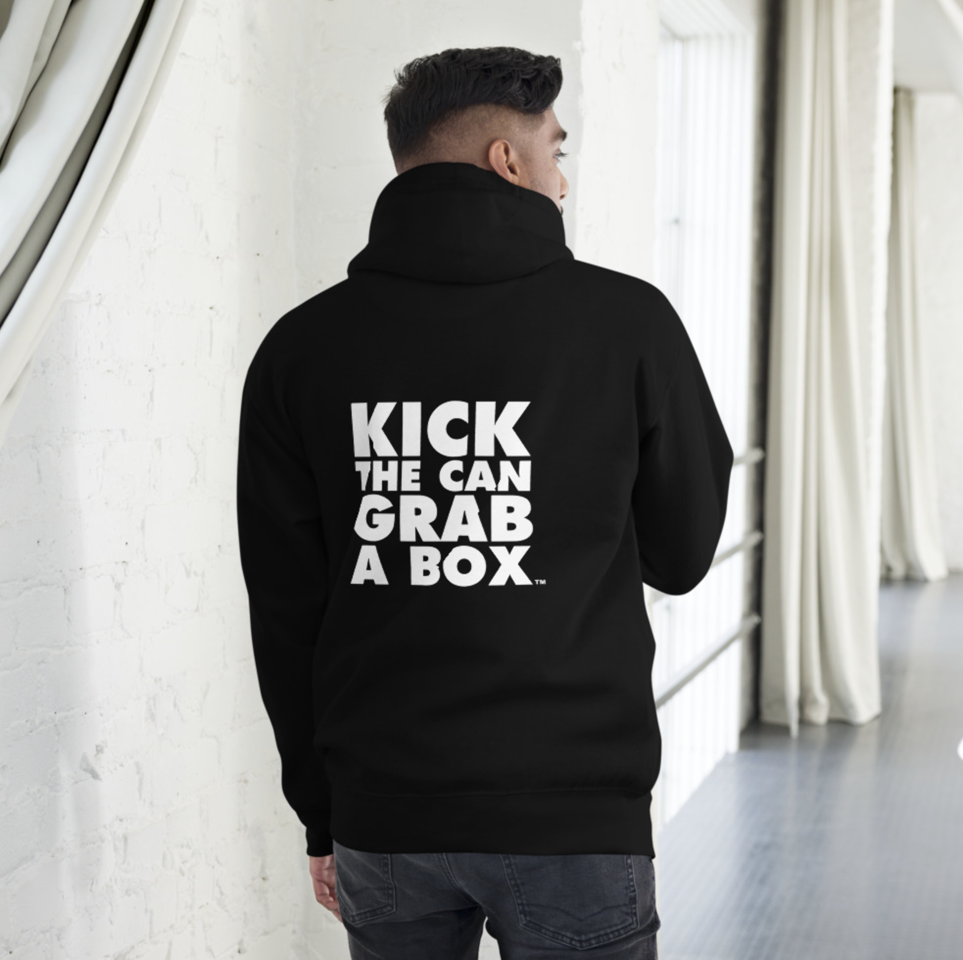 A man wearing a black hoodie with the words "Kick the Can, Grab a Box" printed on the back, standing indoors near a white brick wall and large windows with curtains.