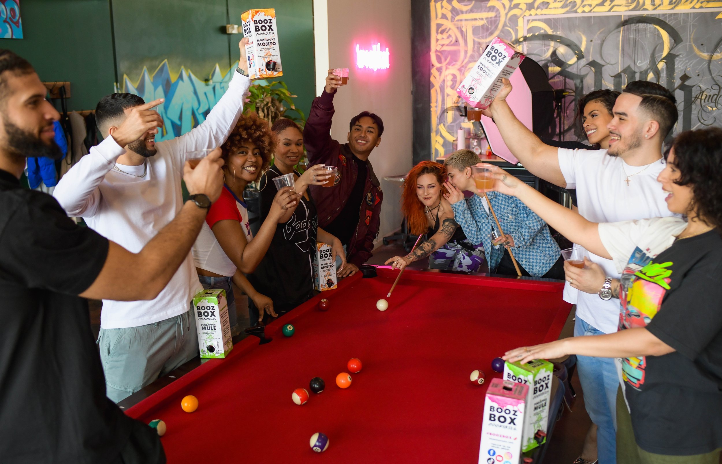 A group of friends celebrating around a pool table, holding drinks and laughing, with some holding boxes of Booz Box cocktails.