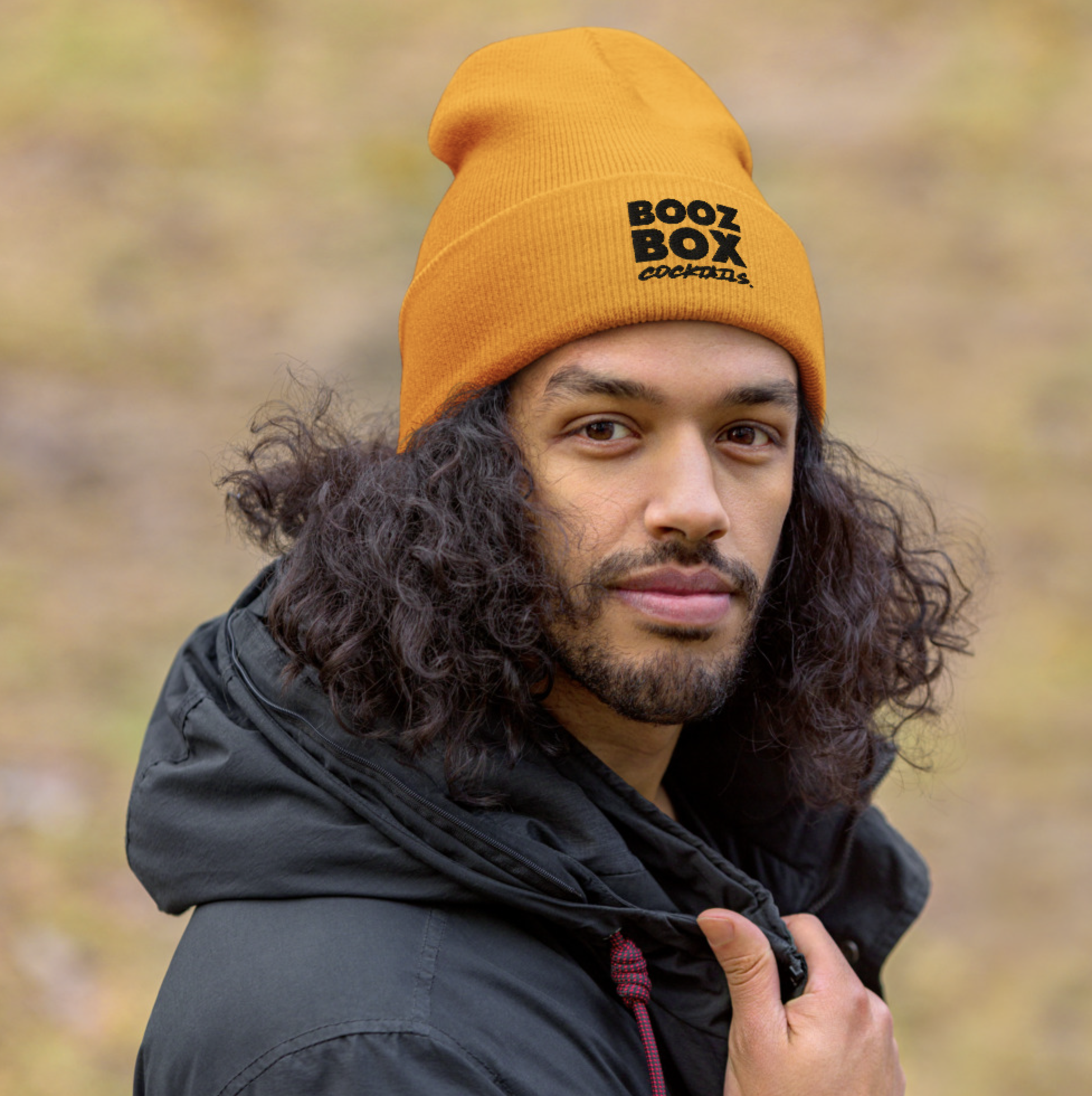 A man with long curly hair and a beard wearing a bright orange beanie that says 'BOOZ BOX COCKTAILS' in black text, and a black jacket, outdoors with blurred natural background.
