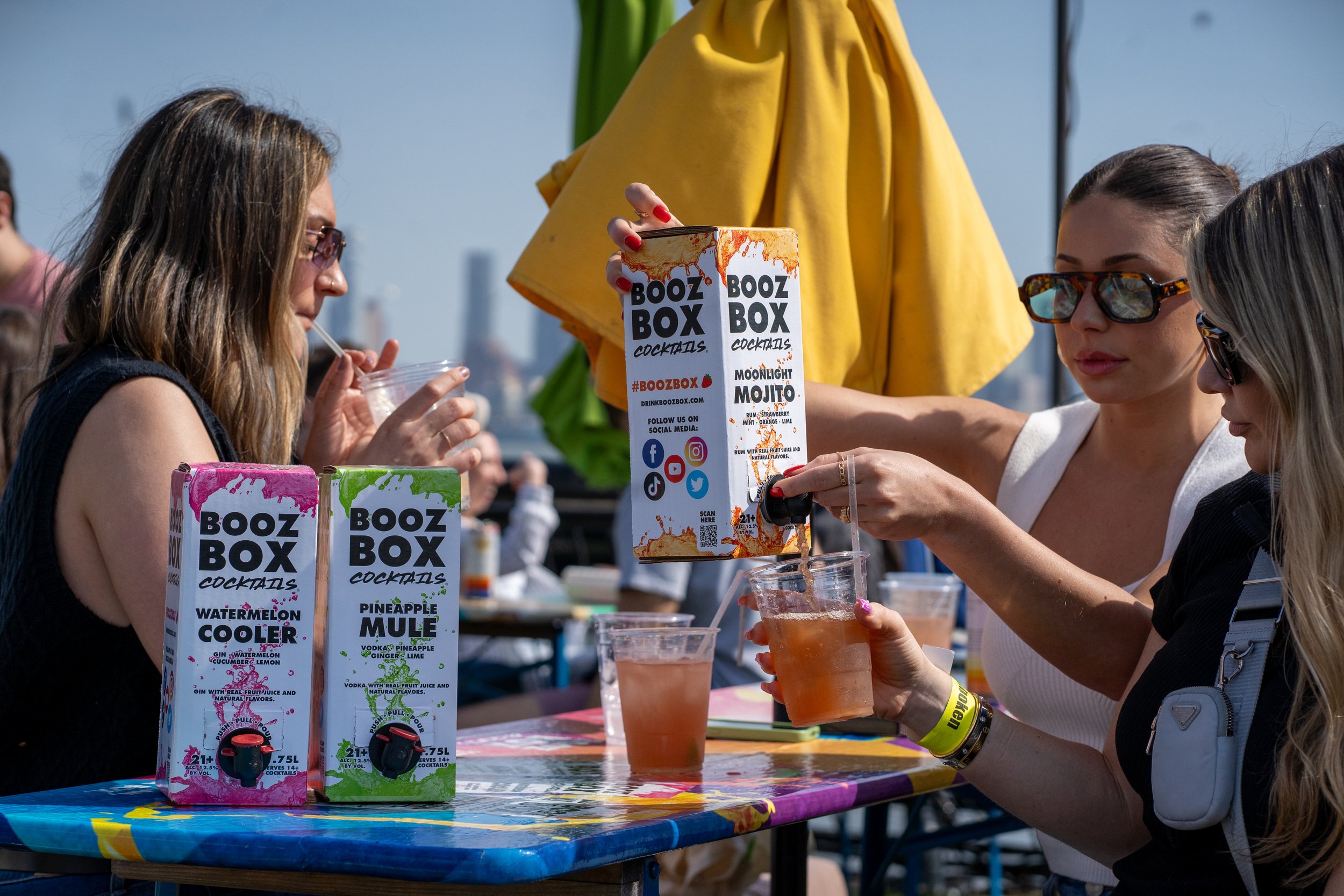 Two women are pouring cocktails from a large box dispenser at an outdoor party or event. There are colorful boxes labeled 'Booz Box Cocktails' on the table, and one woman is drinking from a cup with a straw.