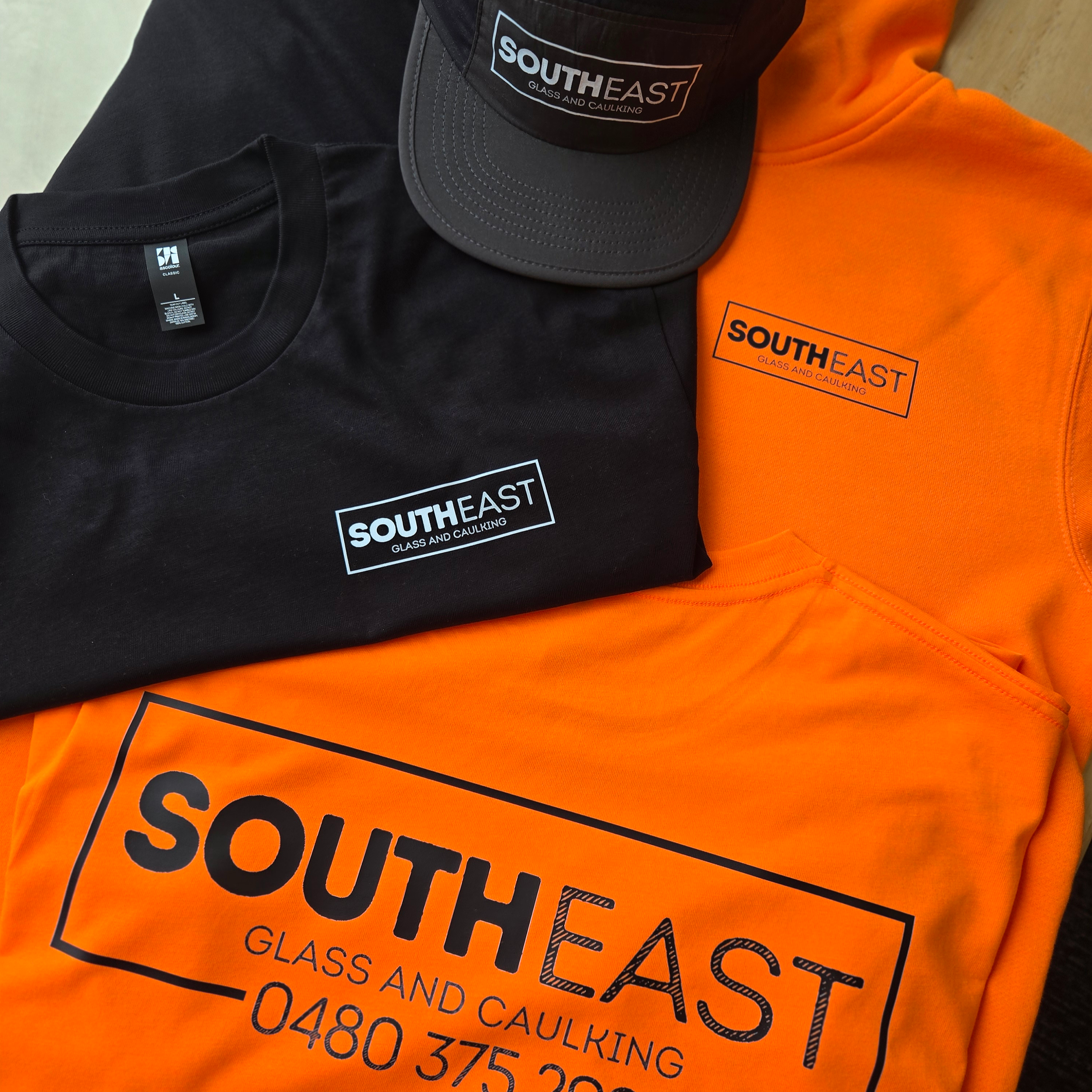 Various South East Glass and Caulking merchandise including a black T-shirt, an orange hi vis tee, an orange hi vis hoodie, and a black cap, all with their new rectangle logo.