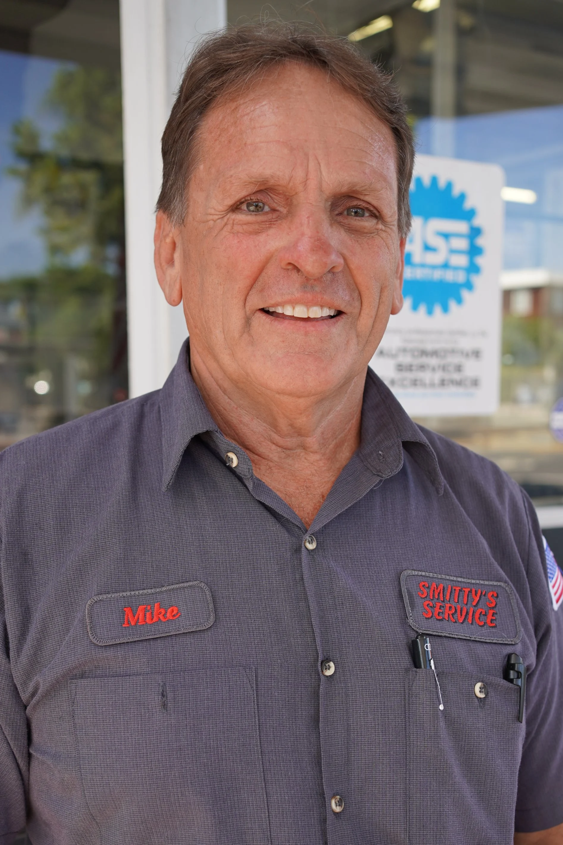 A middle-aged man with short brown hair and blue eyes, smiling, wearing a dark gray work shirt with red embroidered name tag reading 'Mike' and 'Smitty's Service'. He stands outside a building with large glass windows and a sign with a blue gear logo in the background.