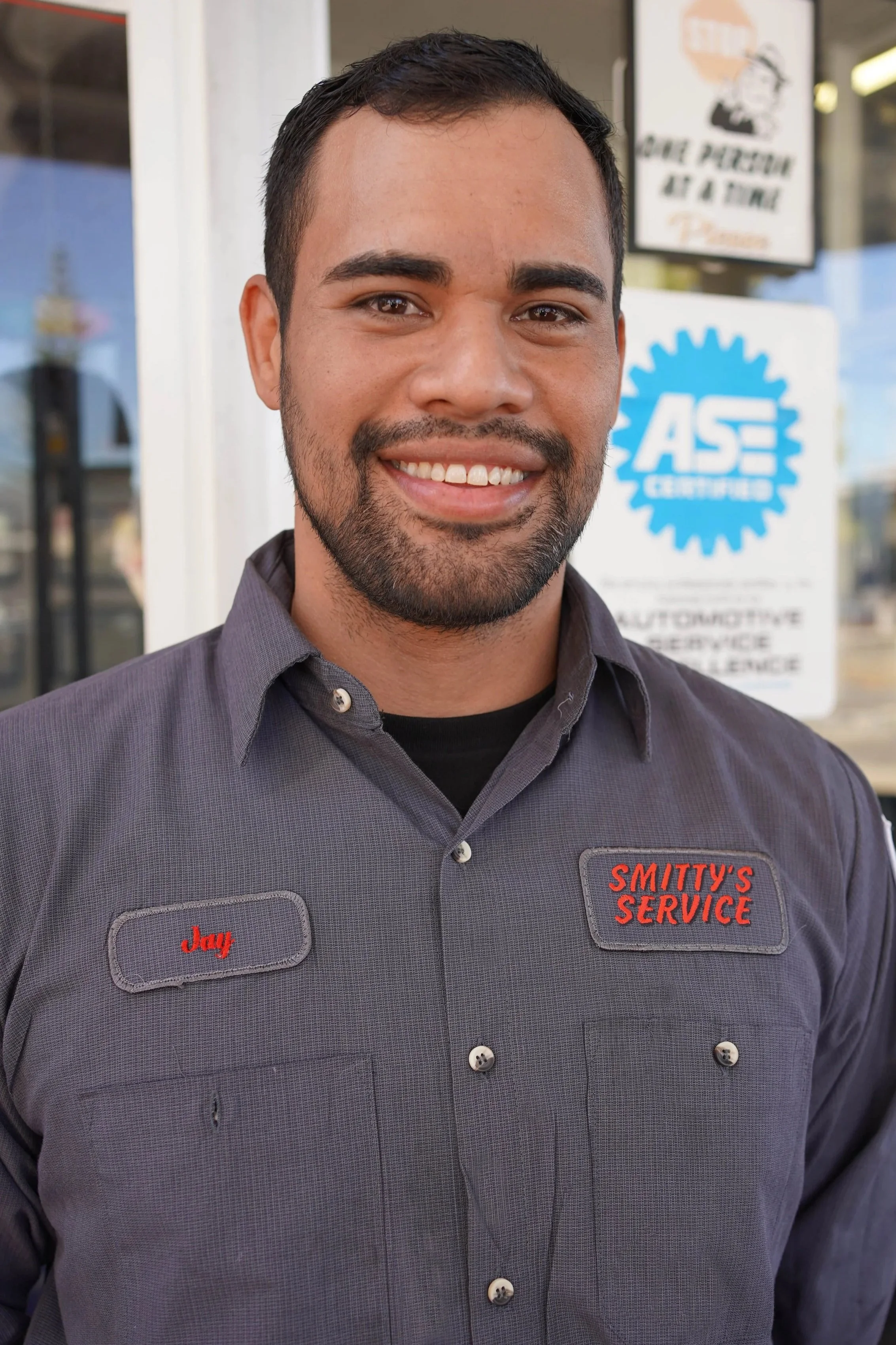 Close-up of a smiling man in a gray work shirt with name tag 'Jay' and company name 'Smitty's Service', standing outside a store with signs in the background.
