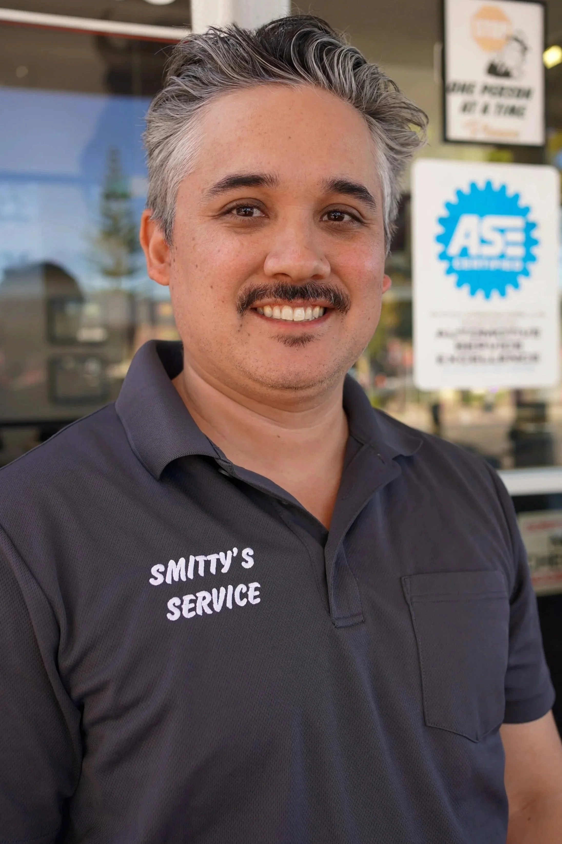 A man with short, gray hair, and a mustache, smiling and wearing a black polo shirt with the words "Smitty's Service" embroidered on it. He is standing outdoors in front of a glass window with signs, including one for ASE certification.