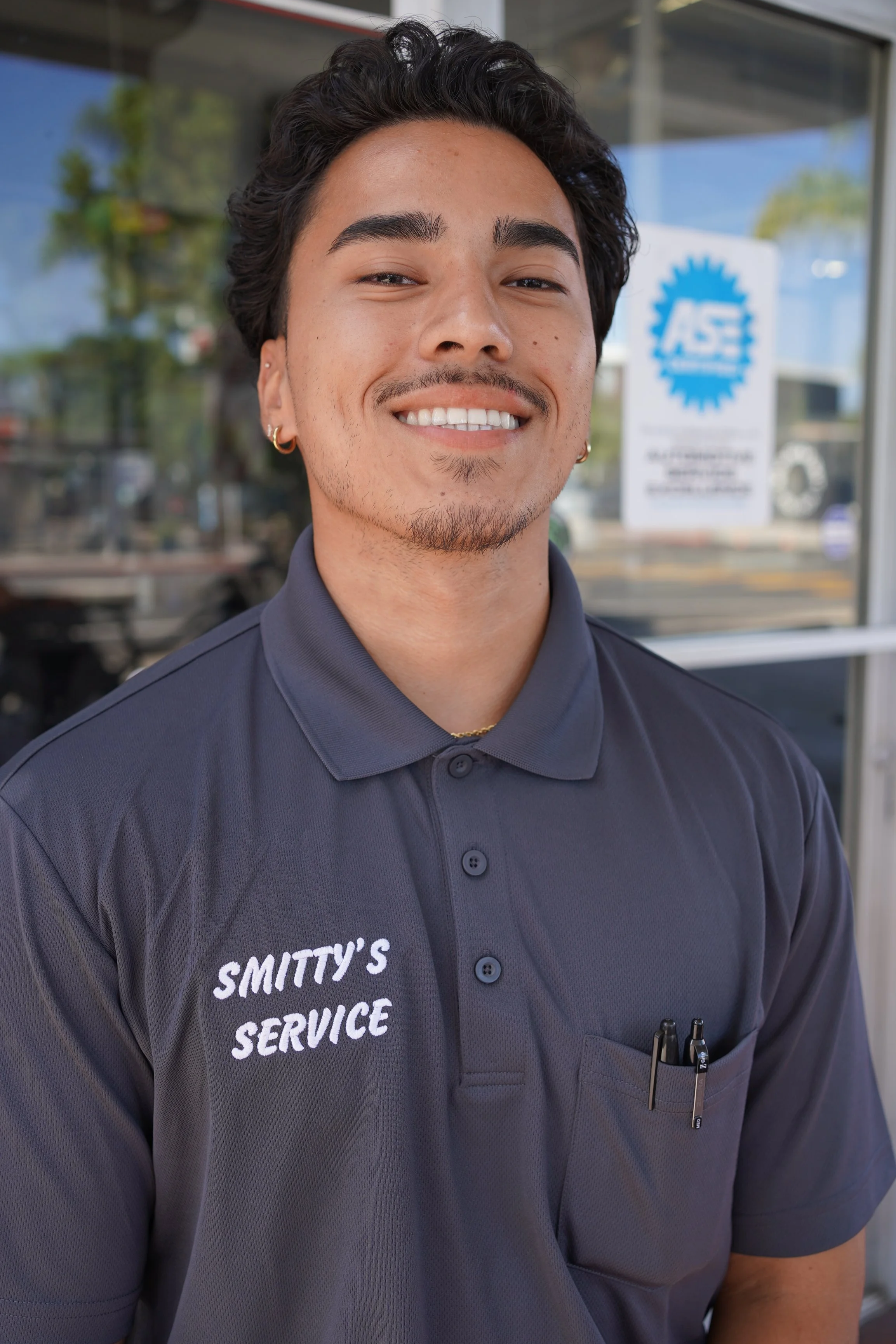A young man with dark, wavy hair, earrings, and a short beard, smiling outdoors in front of a glass window with a faded sign that reads "ASE." He is wearing a dark gray collared shirt with the words "SMITTY'S SERVICE" embroidered on it, and has pens in a pocket.