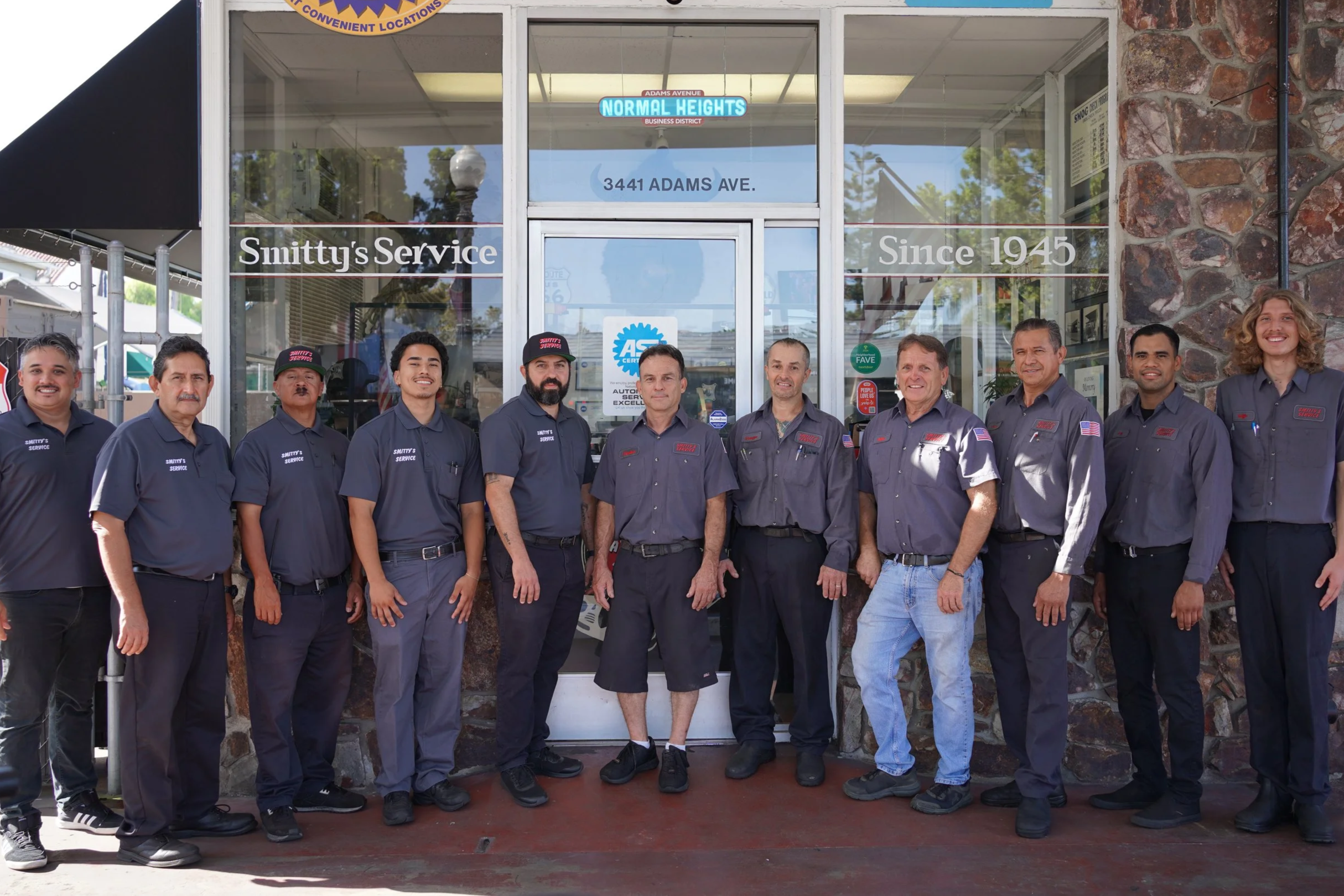 Group of eleven men standing in front of a storefront labeled 'Smitty's Service' with a sign indicating it has been in business since 1945. The men are wearing mechanic uniforms.