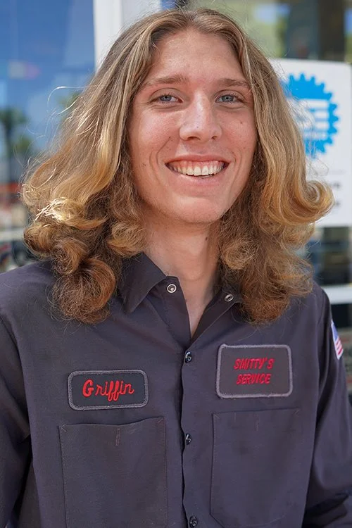 A man with curly blonde hair smiling, wearing a gray work uniform with name tags that say "Griffin" and "Smitty's Service". She is standing outside with a store window and promotional signs in the background.