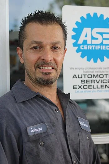 A man wearing a blue work shirt and name tag standing outdoors in front of a sign indicating ASE certification, likely at an automotive service center.