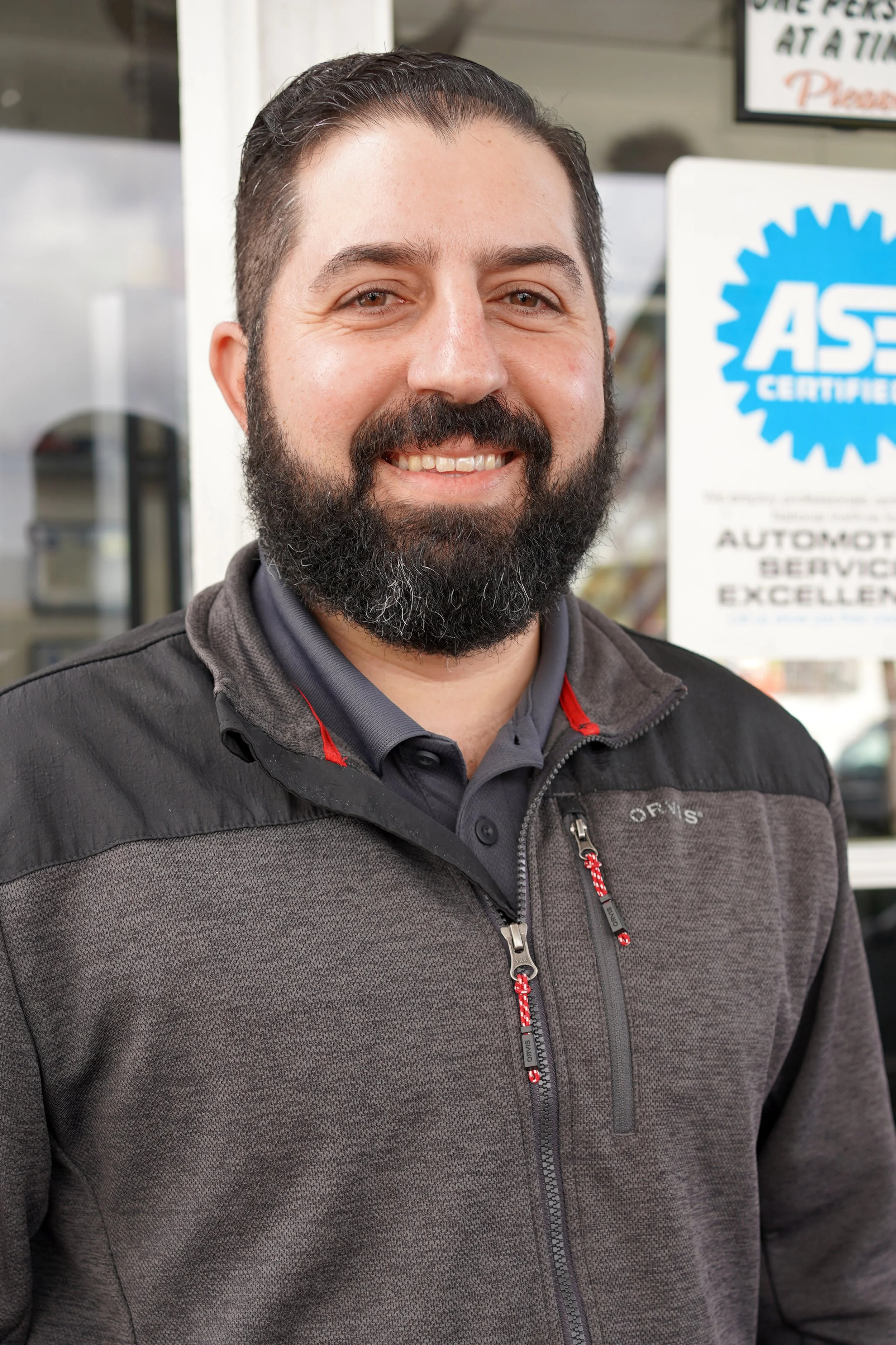A man with a beard and dark hair smiling in front of an automotive service shop, with signs visible in the background.