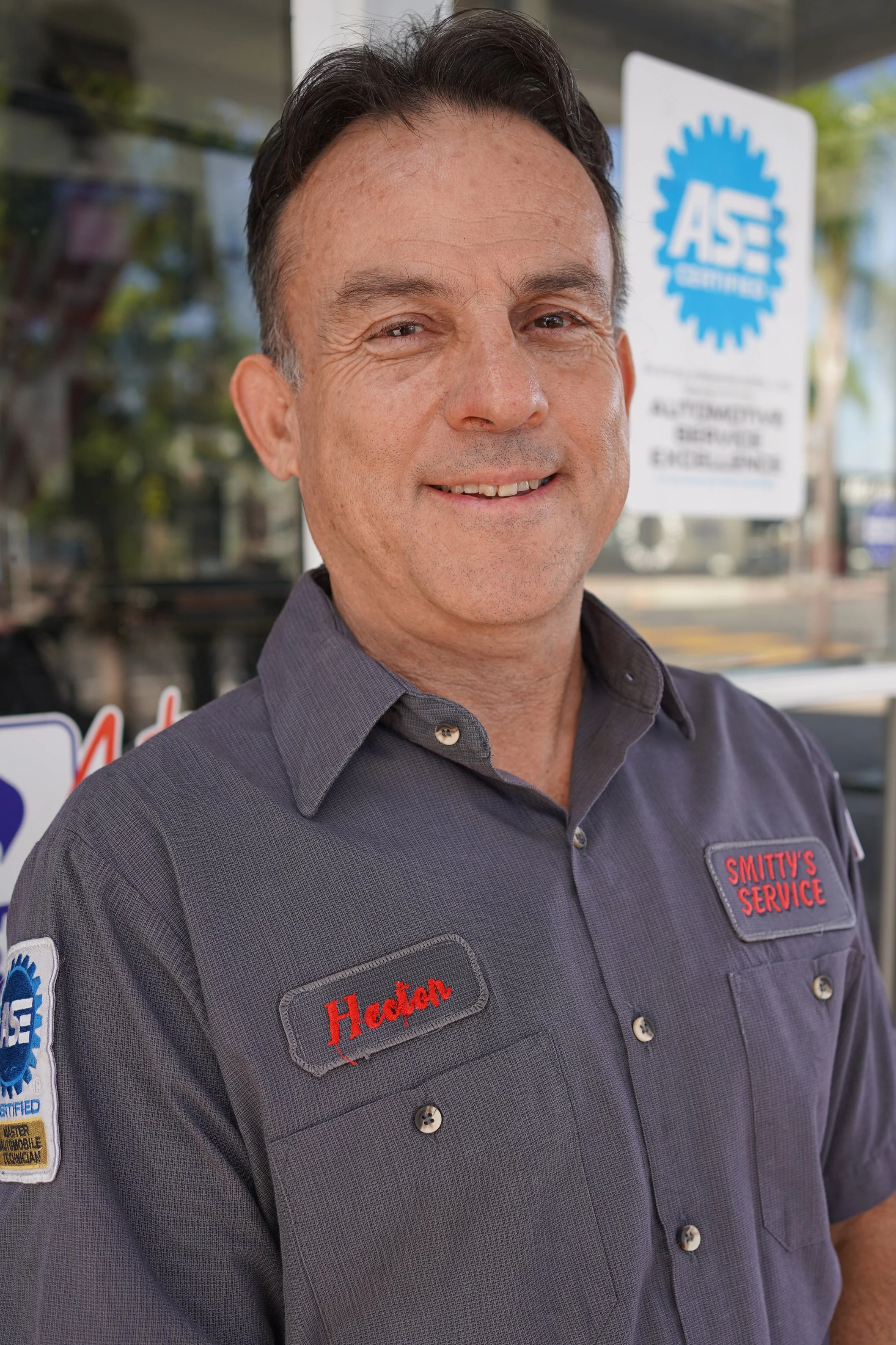 A man smiling, wearing a gray shirt with patches that read "Hedden" and "Smitty's Service," in front of a glass window with a sign that reads "ASE Certified" and workplace information.
