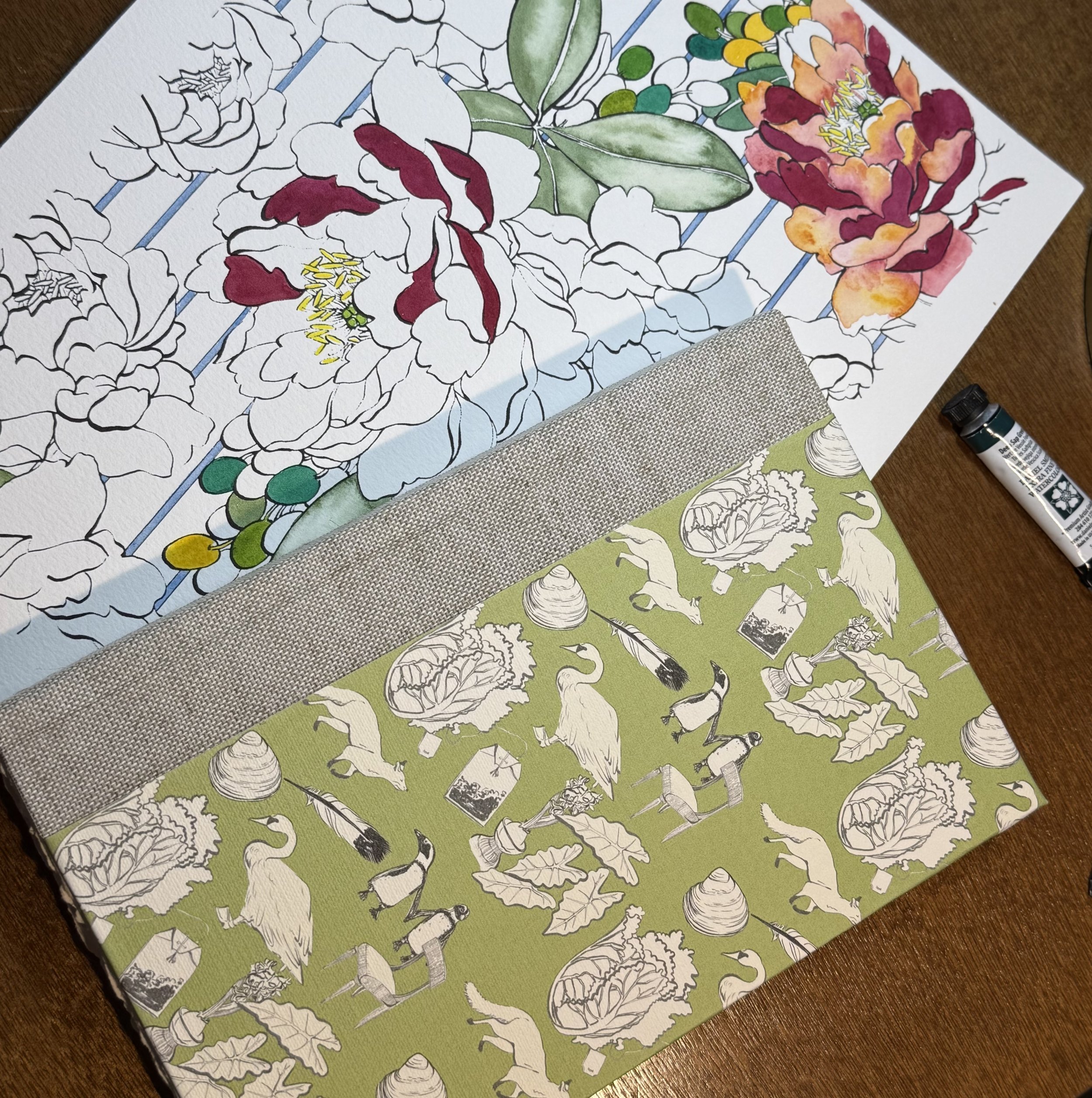 Unfinished coloring pages on a wooden table, one featuring large floral designs with roses and peonies, and another with animal and nature illustrations like cranes, turtles, and trees. A gray fabric strip is placed between the pages, and a black mar