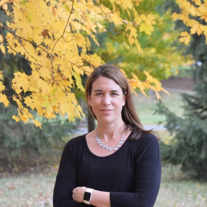 Carolina Fabbi with  brown hair, wearing a black top and a diamond necklace, standing outdoors beneath yellow autumn leaves, with a background of trees and grass.
