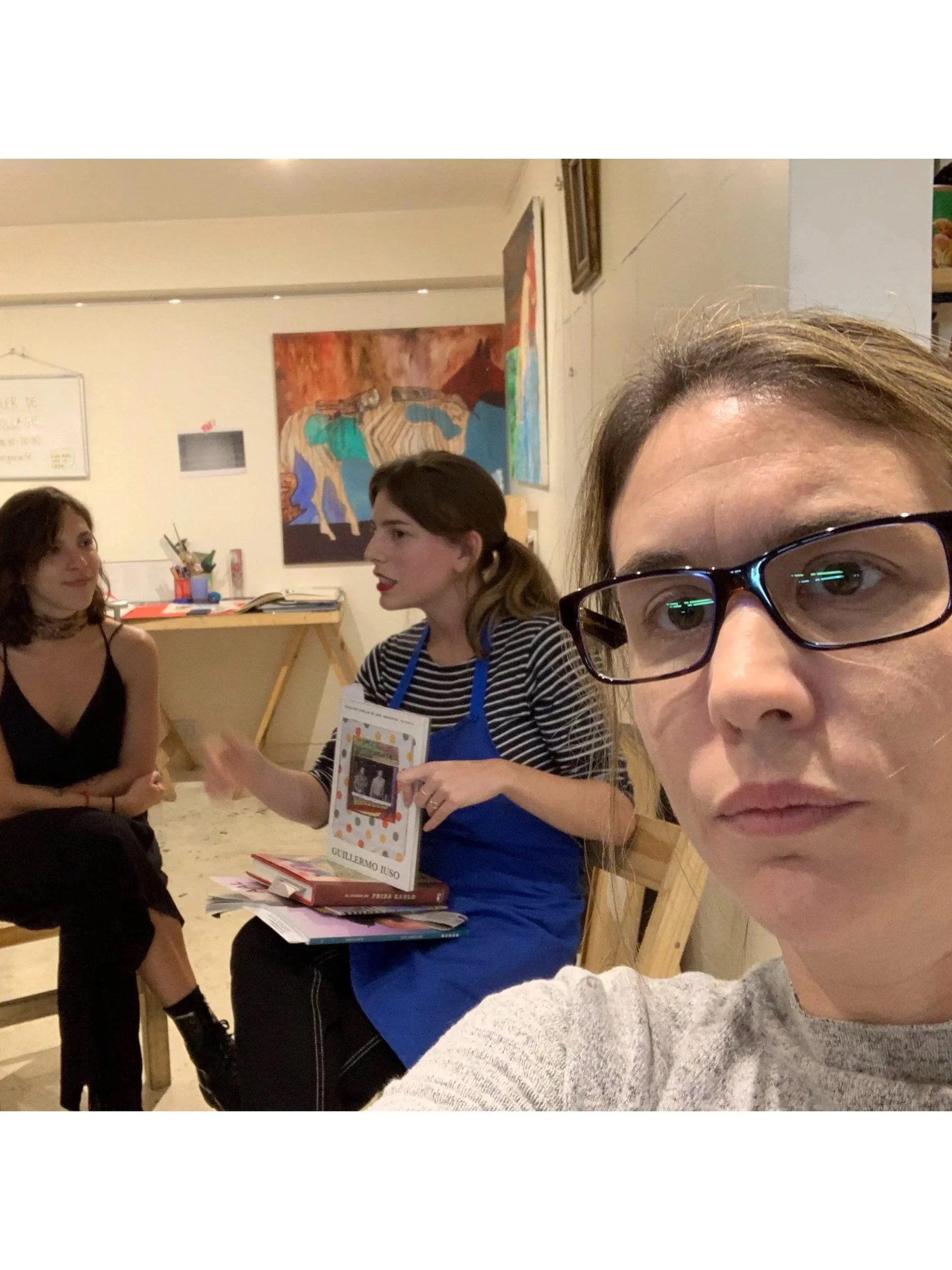 A woman with glasses taking a selfie in a room where two other women are sitting and talking. One woman is wearing a black dress and the other is wearing a striped shirt with a blue apron, holding a book.