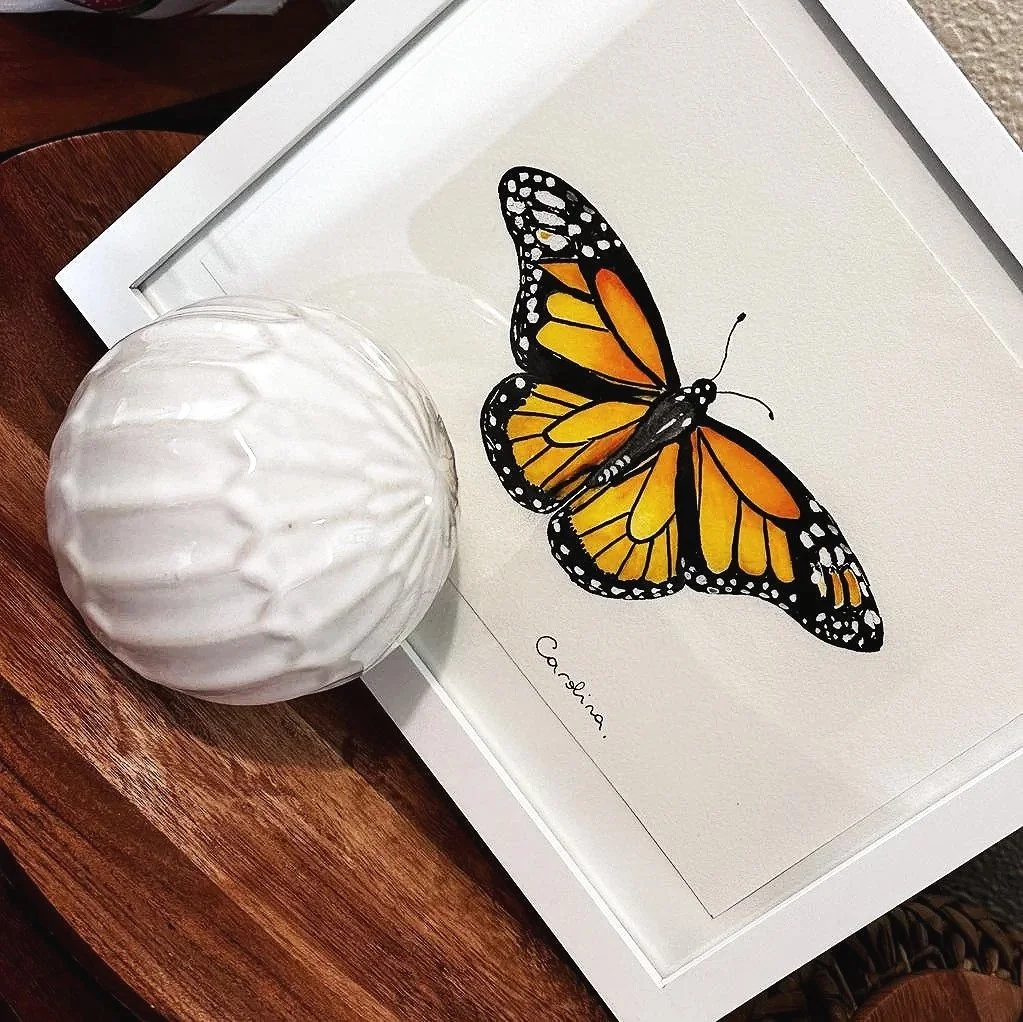 Watercolor painting of a monarch butterfly with orange and black wings, positioned on a beige background. Next to the painting is a white, textured ceramic vase with a round shape and ridged surface, resting on a wooden surface.