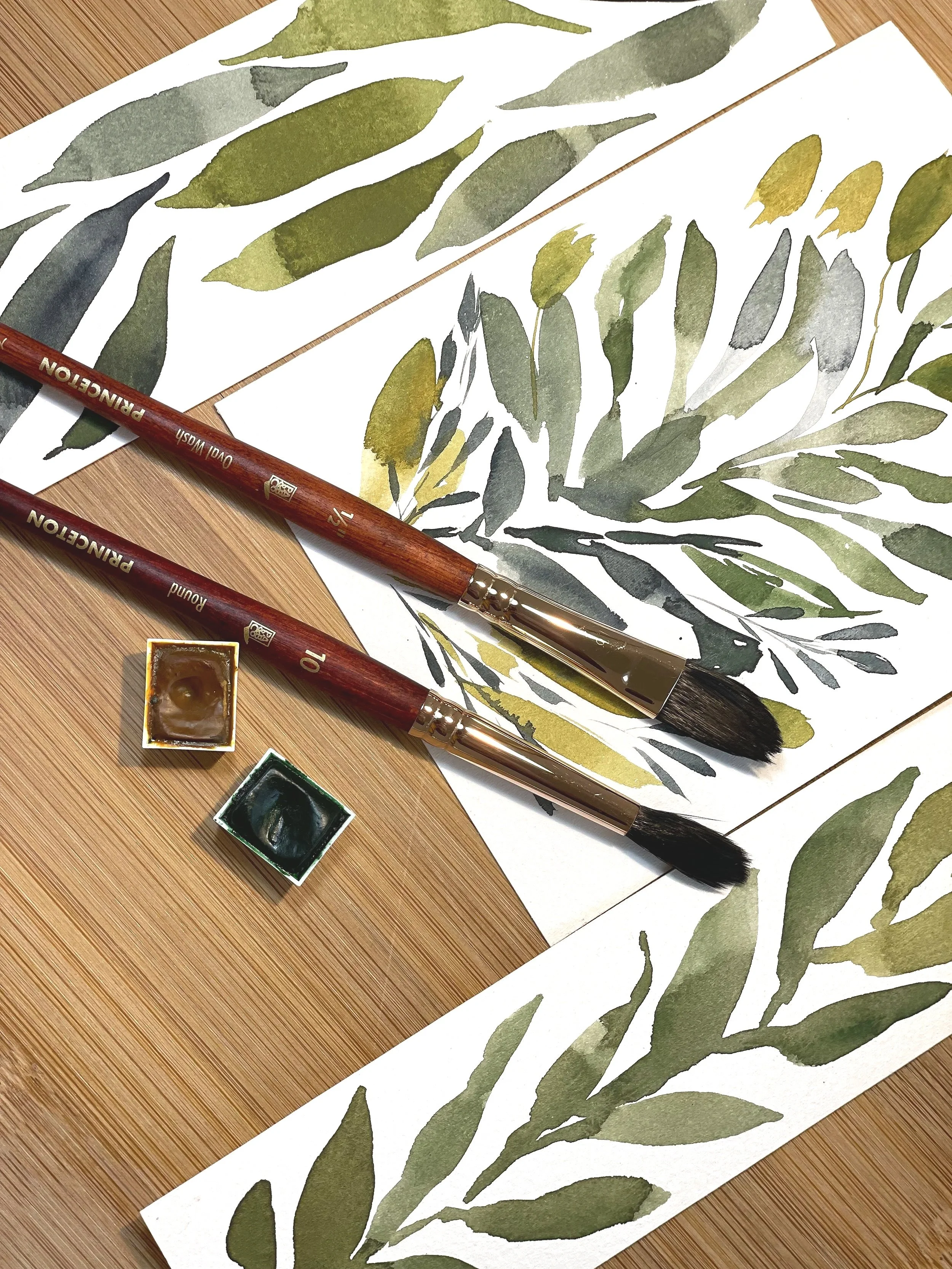 Watercolor paintings of green leafy branches on white paper, two round paint containers with green and brown paint, and two paintbrushes with wooden handles and black bristles on a wooden surface.