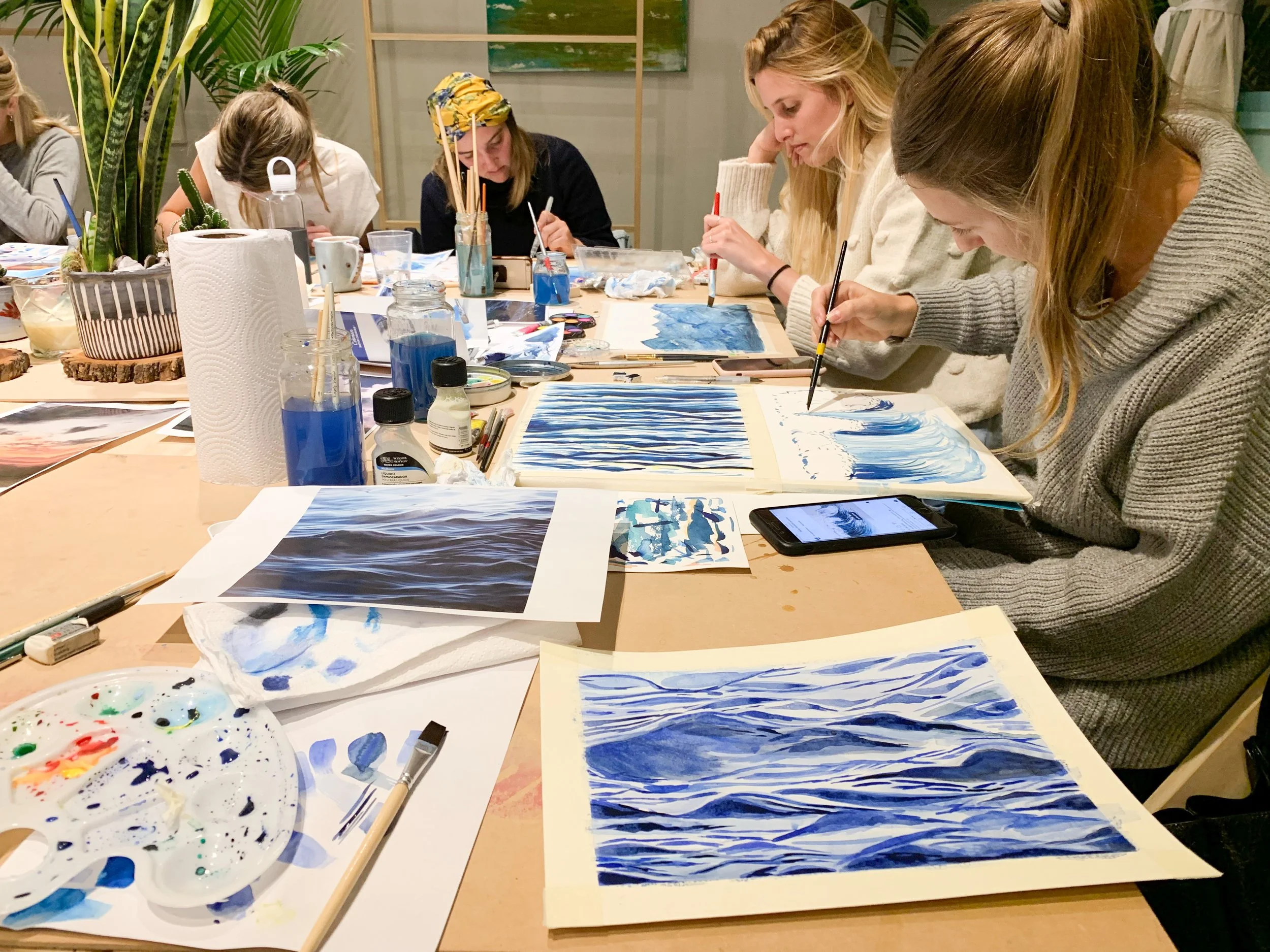 People participating in a watercolor painting class, painting blue and white ocean scenes at a large table covered with art supplies.