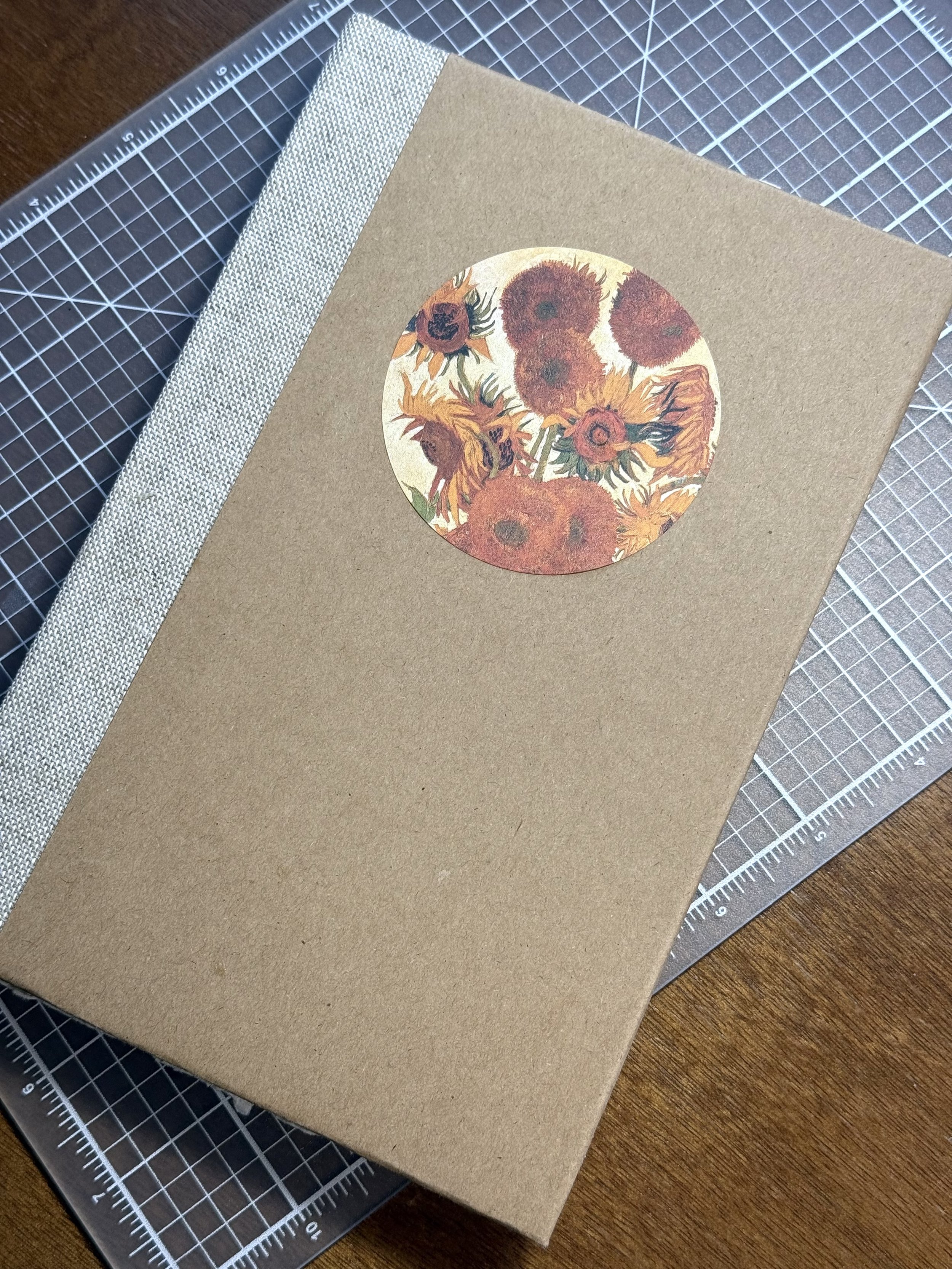 Brown craft paper notebook with a circular sticker of sunflowers on the cover, placed on a cutting mat with rulers.