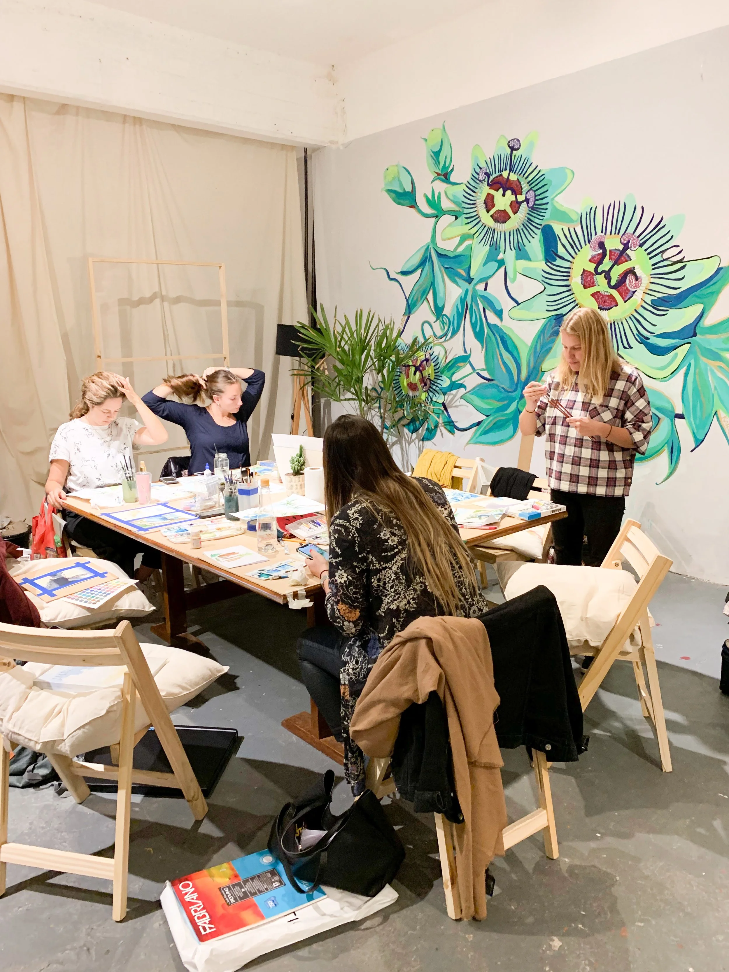 A group of women working at a table in a creative or art studio space, with a large floral mural on the wall behind them.