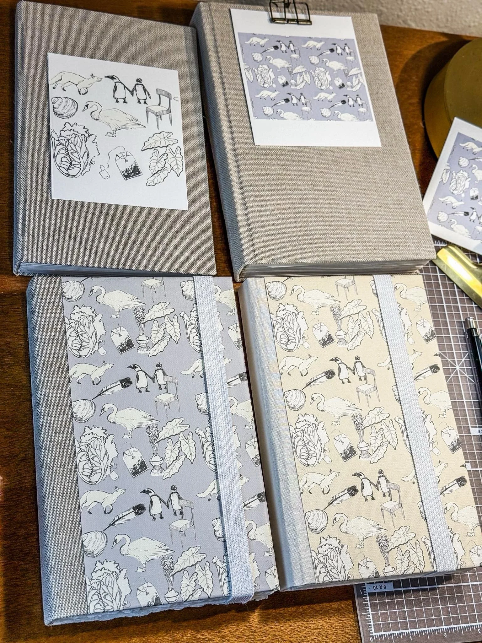 Four fabric-bound notebooks and papers on a wooden table, some with sketches and patterned designs of animals and plants, along with a cutting mat and pen.