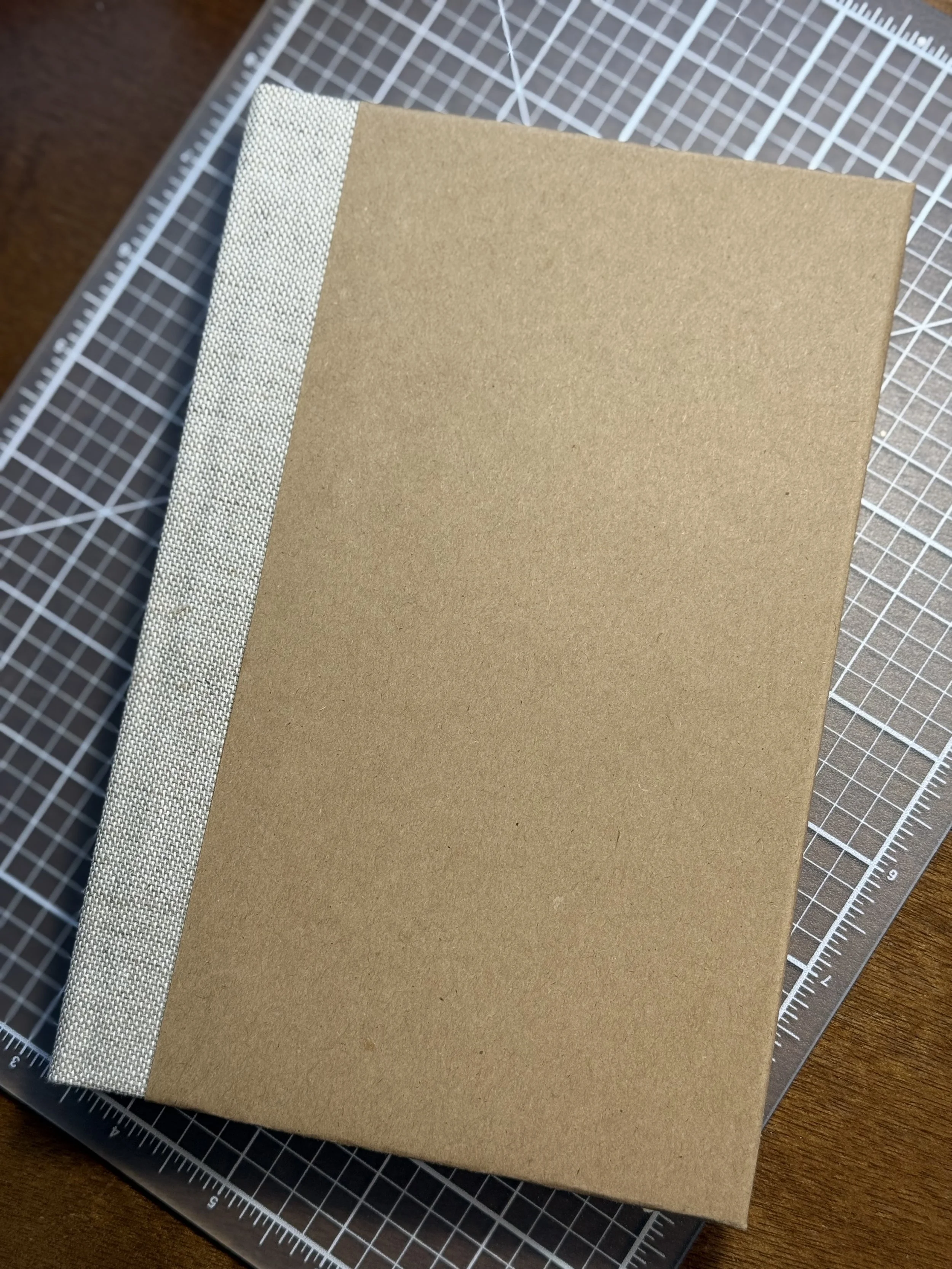 A brown, hardcover book with a light fabric spine, placed on a grid cutting mat on a wooden surface.