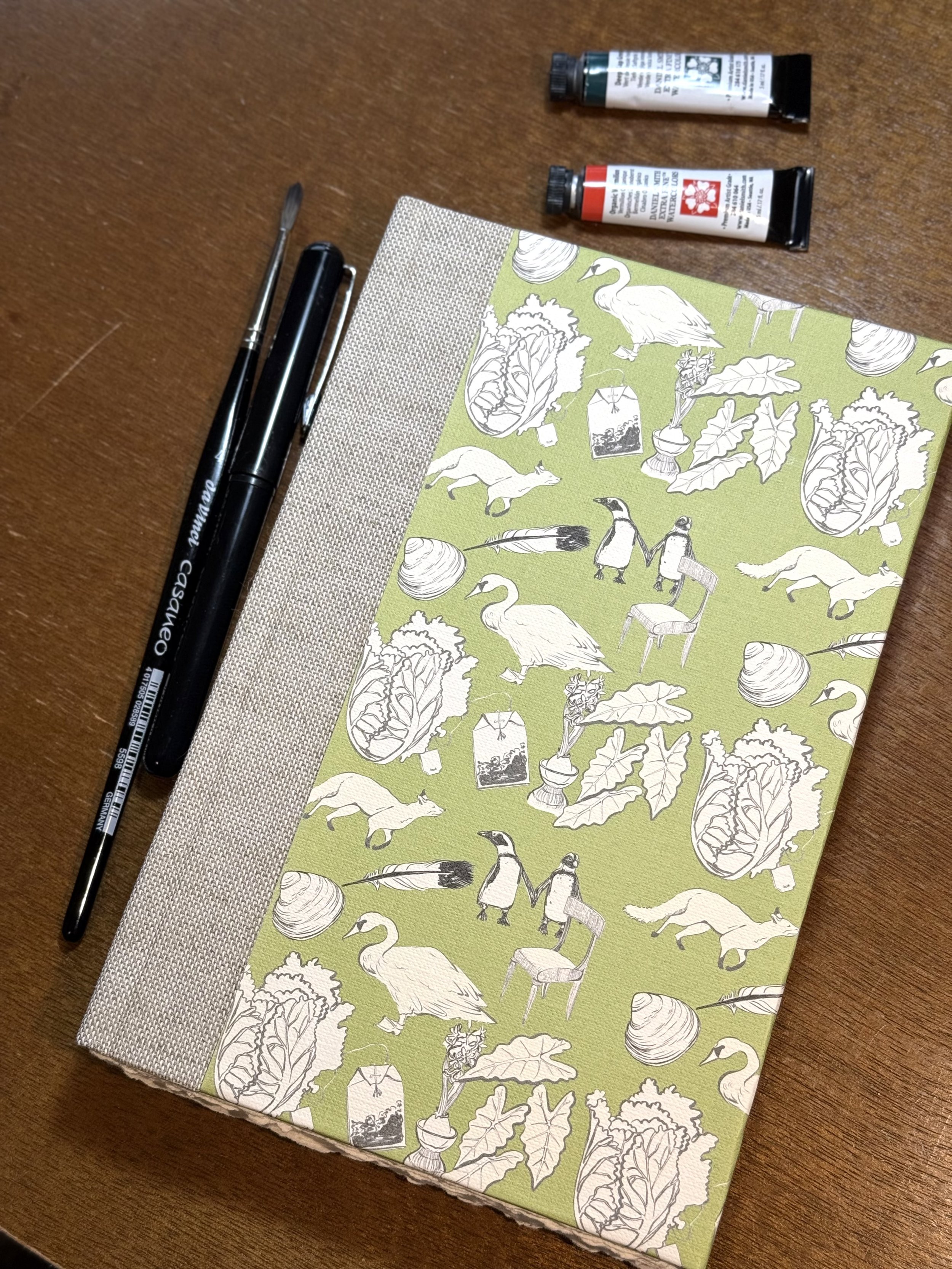A notebook with a green cover decorated with illustrations of swans, foxes, igloos, leafy vegetables, and potted plants, placed on a wooden surface along with a black pen, a small flathead screwdriver, and two tubes of paint.