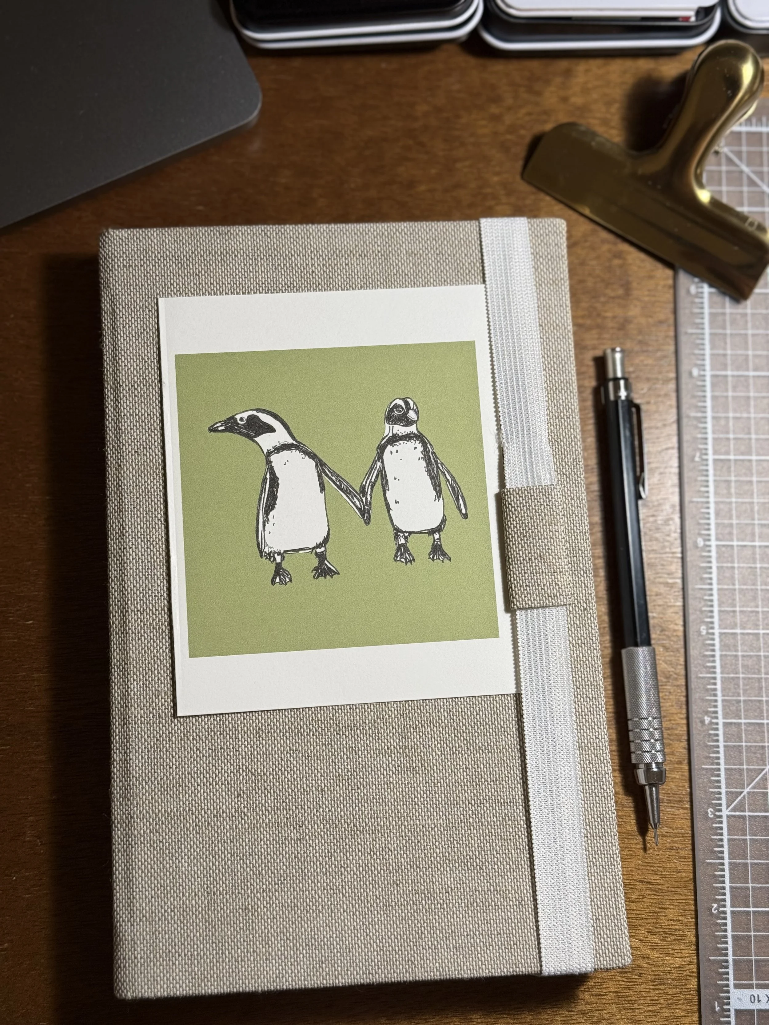Close-up of a beige fabric-covered notebook with a photo of two penguins holding hands on the cover, placed on a wooden desk along with a black pen, a metal thimble, a brass hammer, and a ruler.