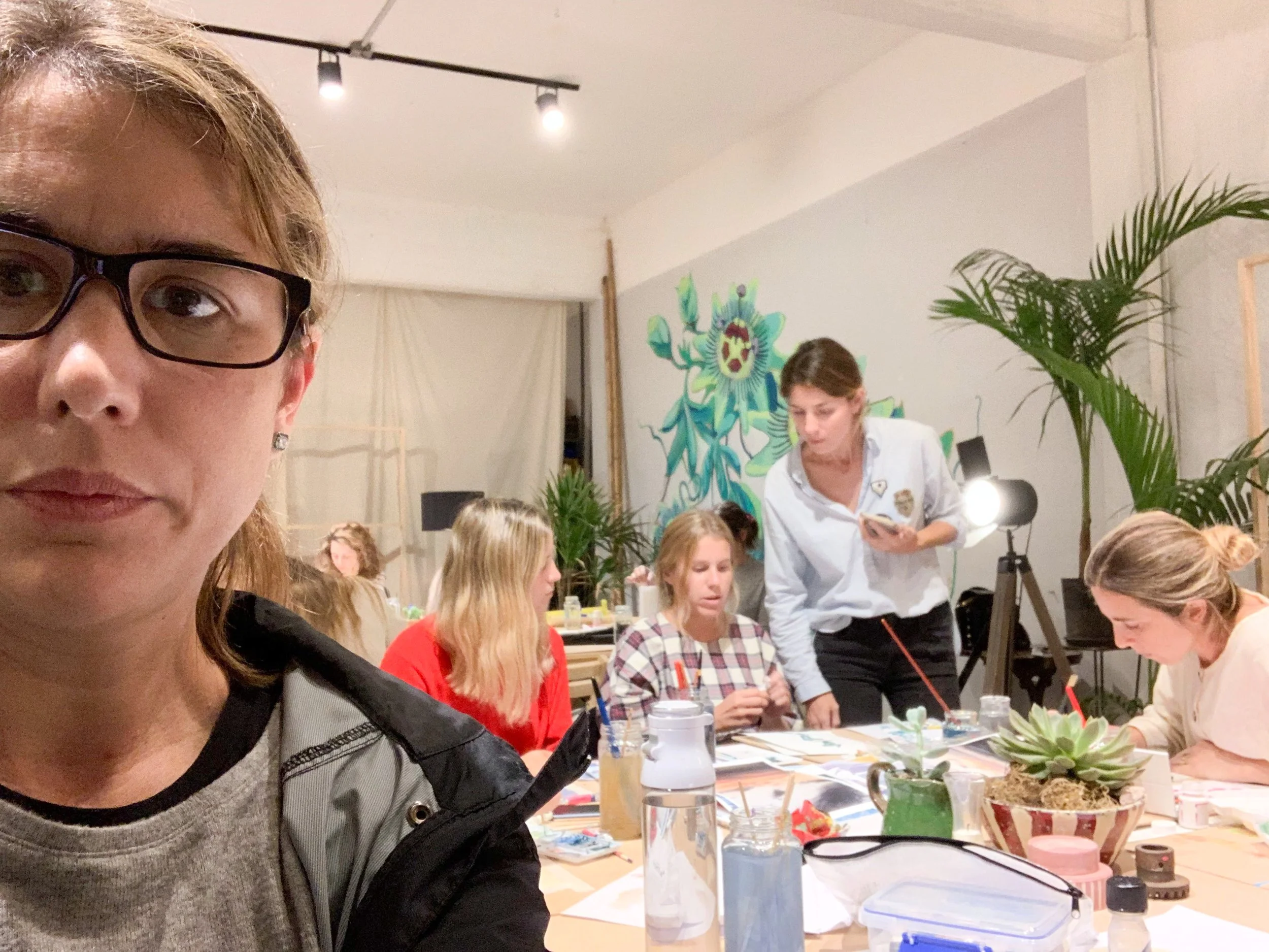 A woman in the foreground taking a selfie, with a group of women and a man in the background working together at a table in a creative space with plants and colorful wall art.