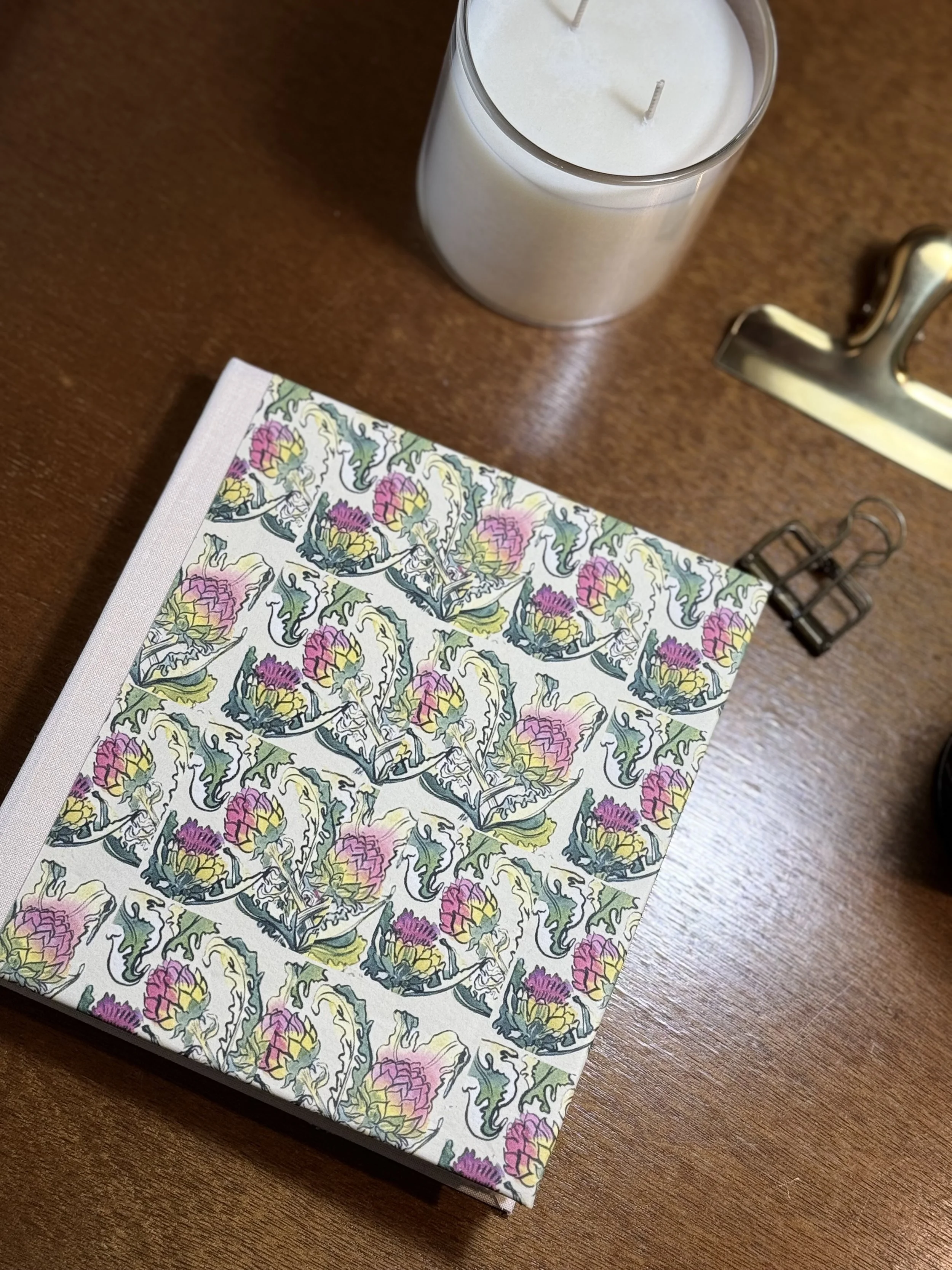 A closed book with a floral pattern cover featuring pink, yellow, and green colors, placed on a wooden surface. A white candle in a glass container is nearby, with part of a metallic object and a black binder clip visible.
