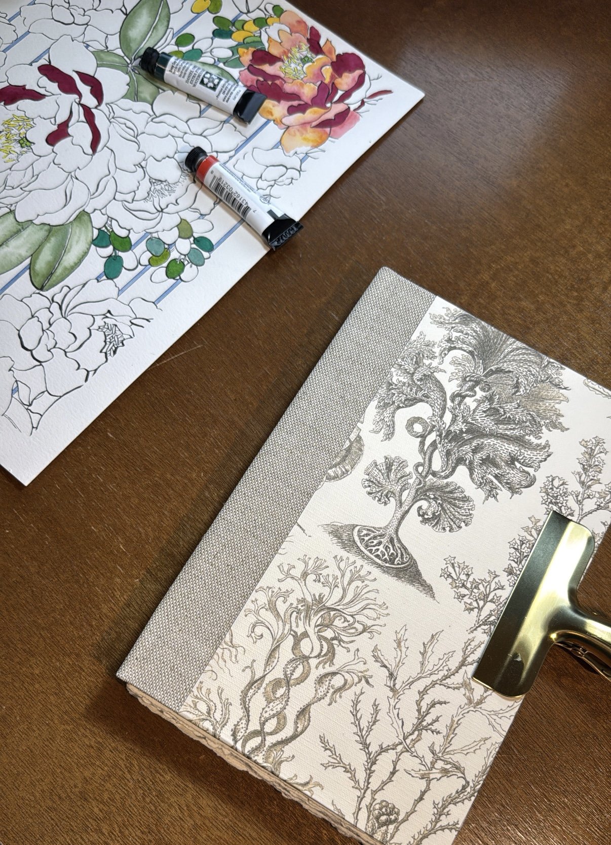 A wooden table with a coloring page featuring a large flower and leaves, colored partially with paints. There are two open paint tubes on the coloring page and a closed, decorative book with a botanical illustration of trees and plants on the cover.