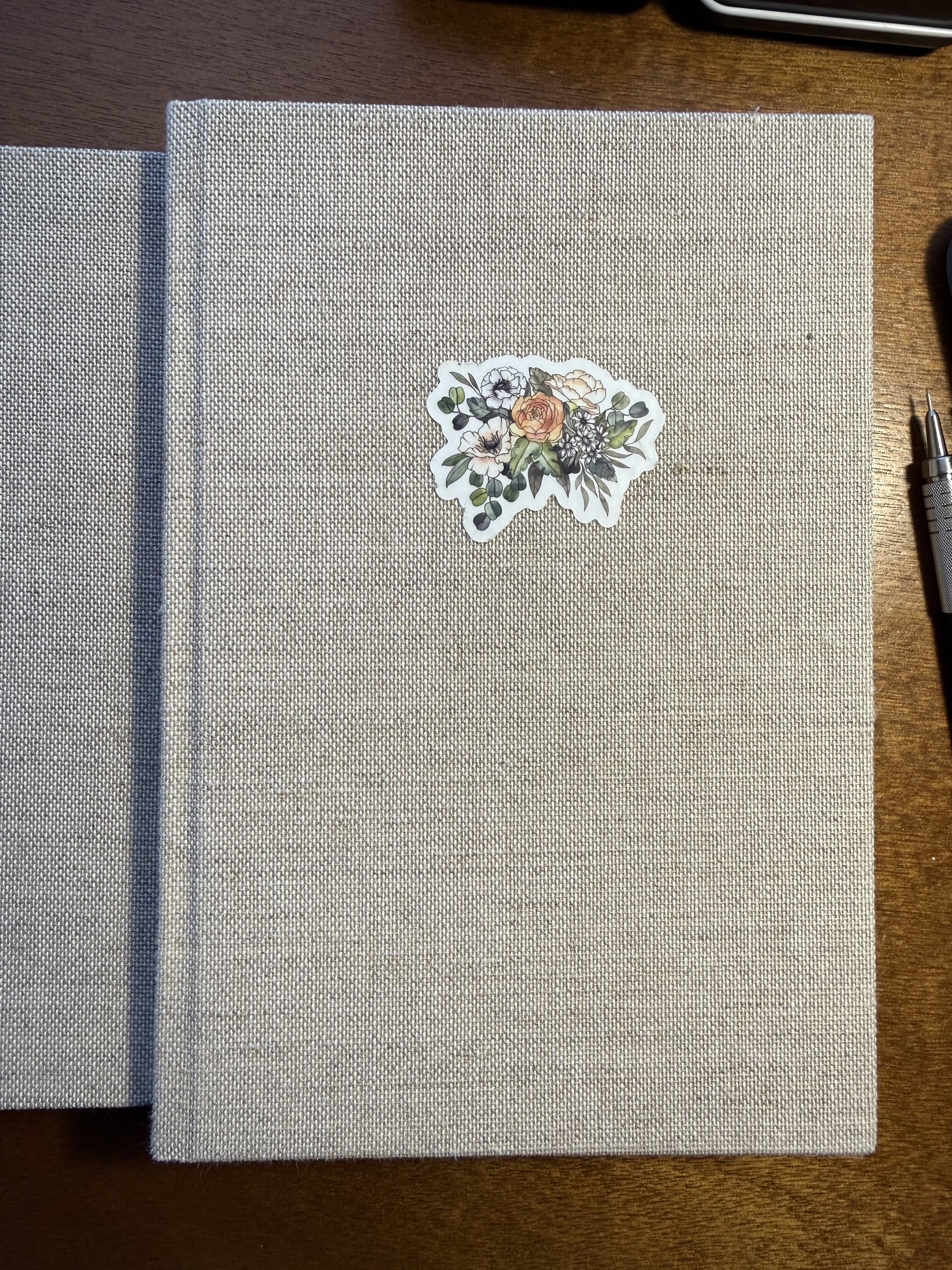 A closed beige fabric-covered notebook with a floral sticker on the cover, placed on a wooden surface.