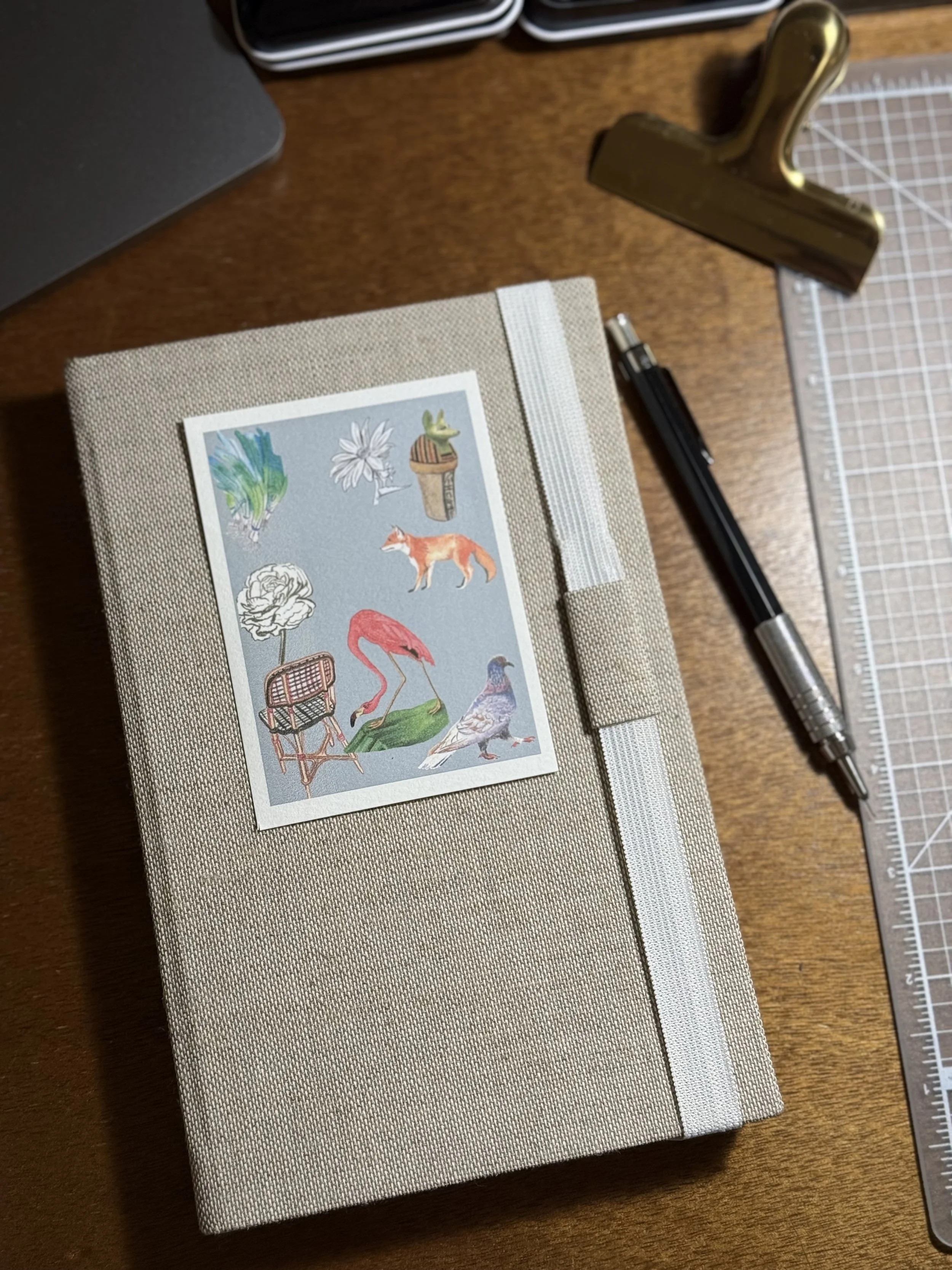 A beige fabric-covered journal with a sticker of various colorful animals and plants on the front, placed on a wooden desk. Nearby are a black pen, a gold binder clip, and a clear grid cutting mat.