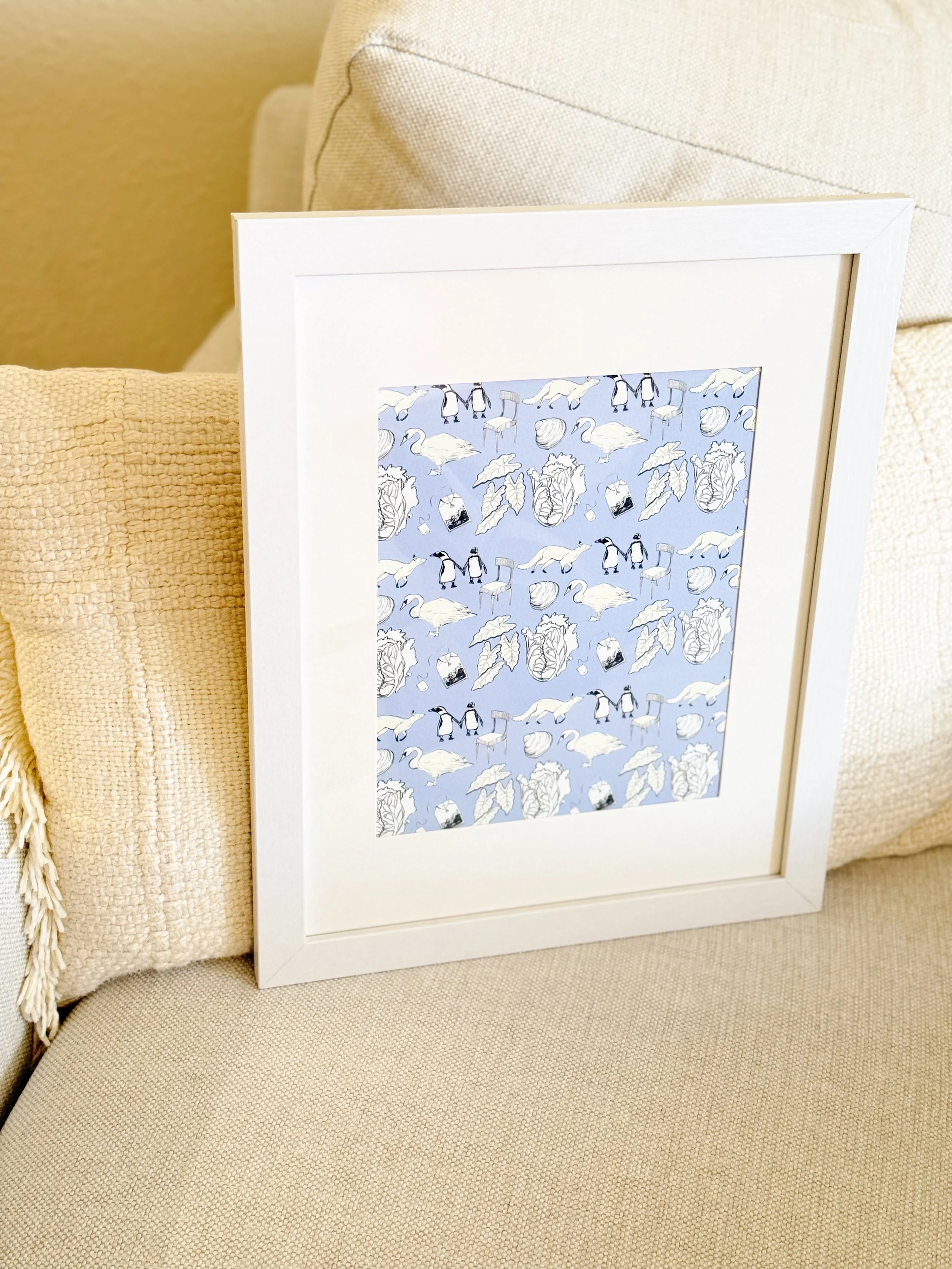 Framed artwork featuring penguins, swans, seashells, leaves, and abstract shapes against a light blue background, leaning on a beige couch with textured pillows.