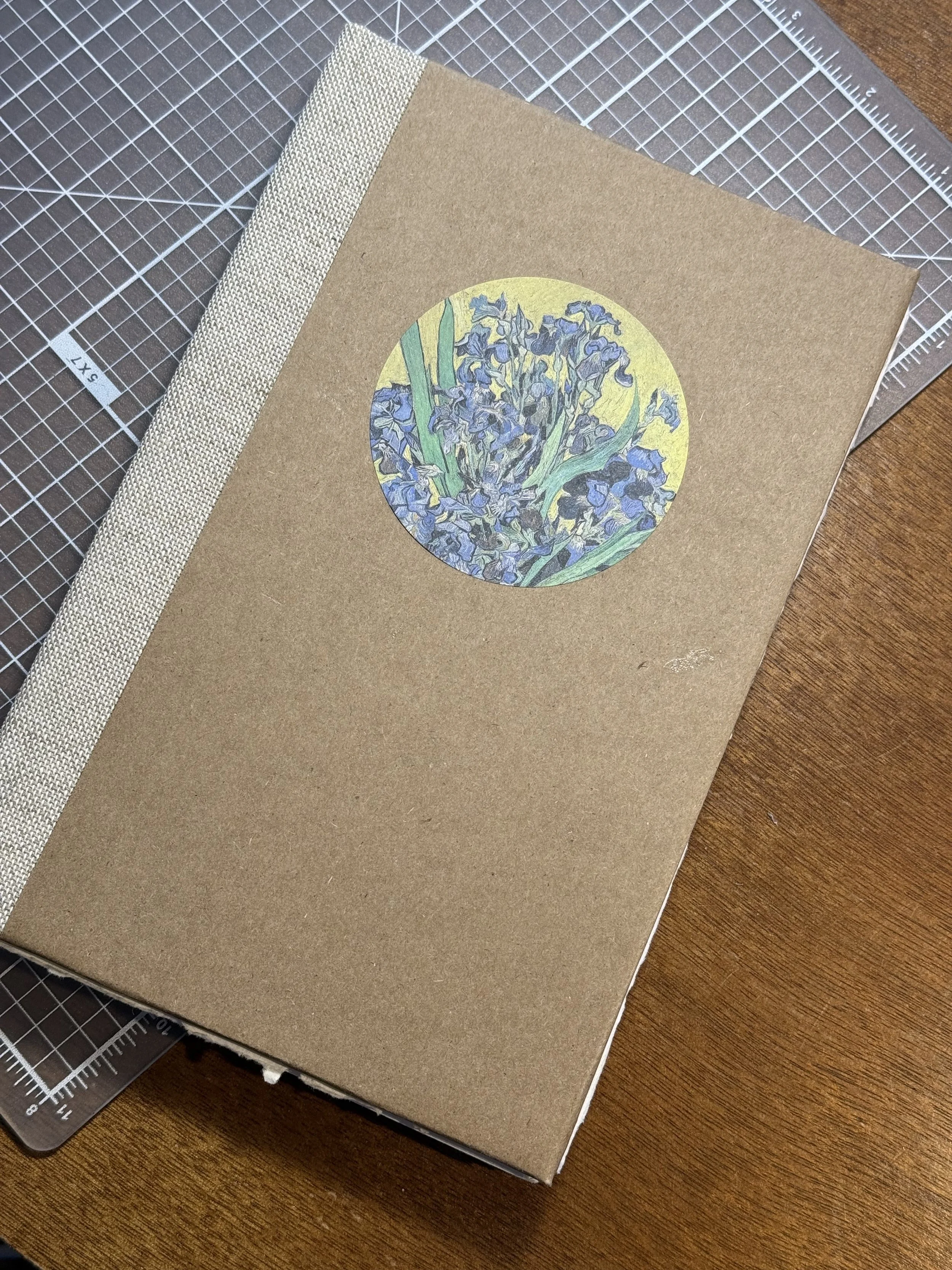 A brown hardcover book with a circular illustration of blue irises on the cover, placed on a cutting mat on a wooden surface.