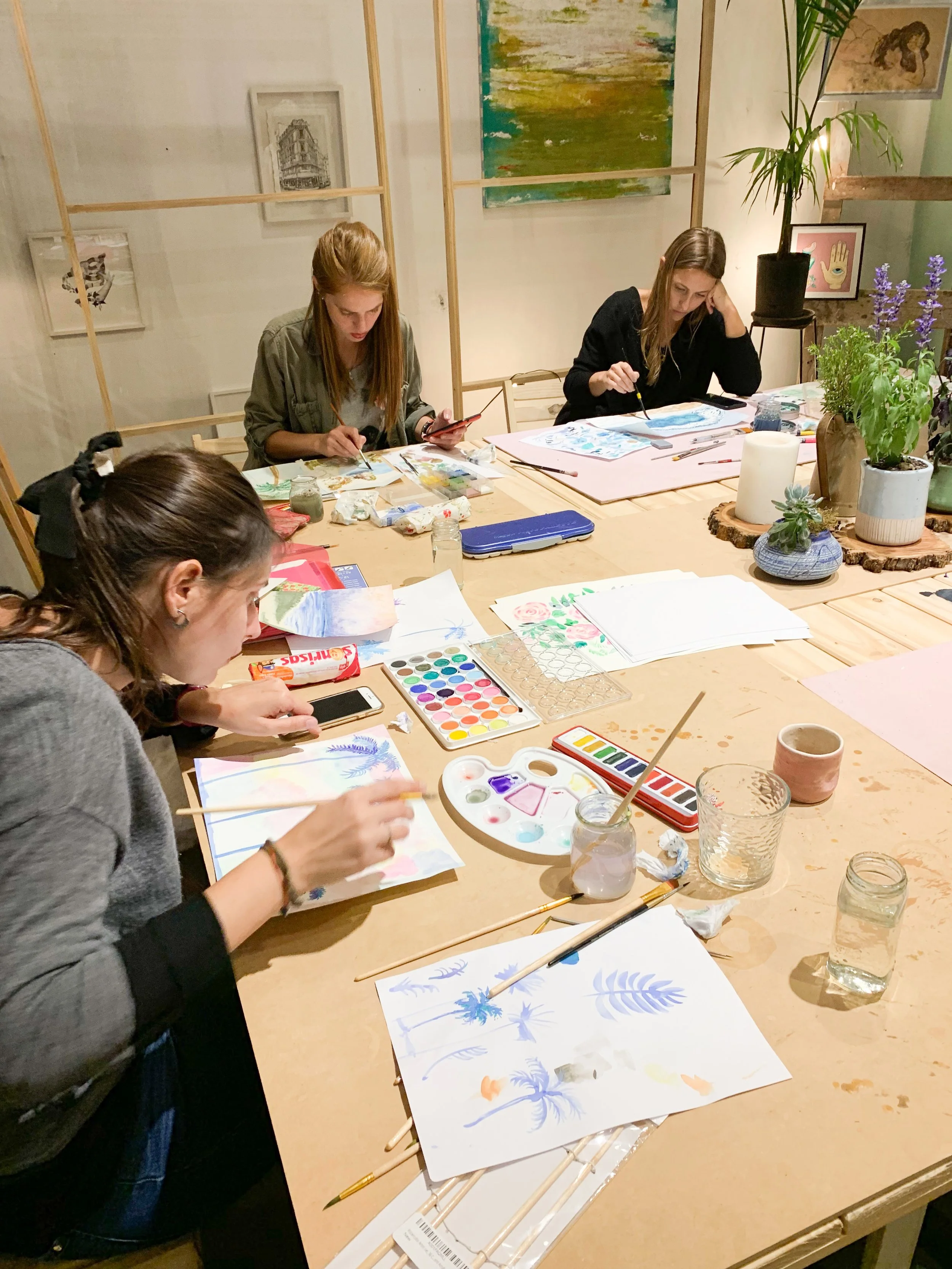 People painting watercolor art at a table with paints, brushes, and paper in an art studio.