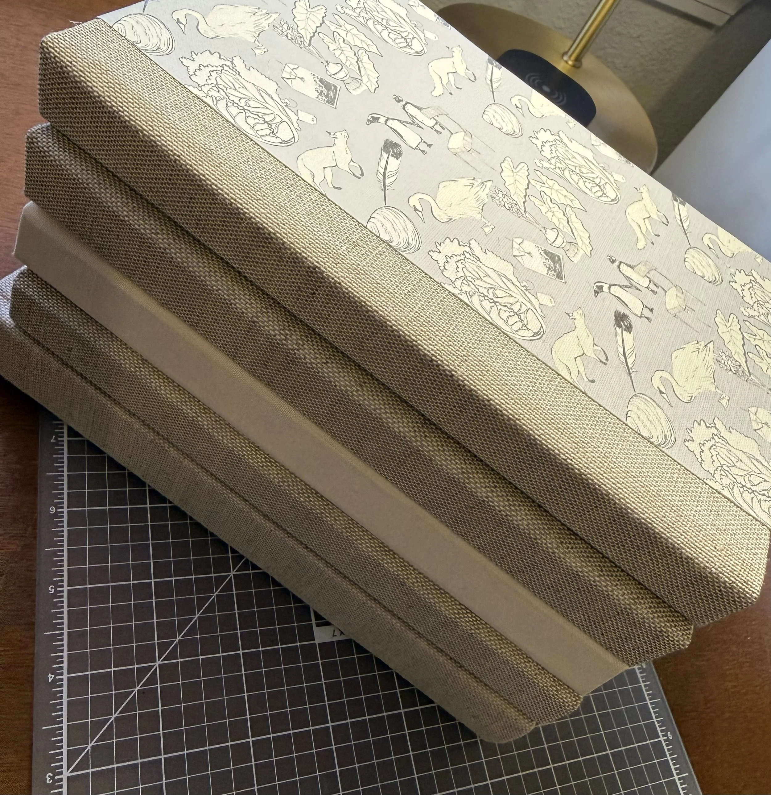 A stack of six fabric-covered books or boxes with a textured beige fabric, placed on a cutting mat with measurement grid, next to a wooden table and a lamp with a round wooden base.