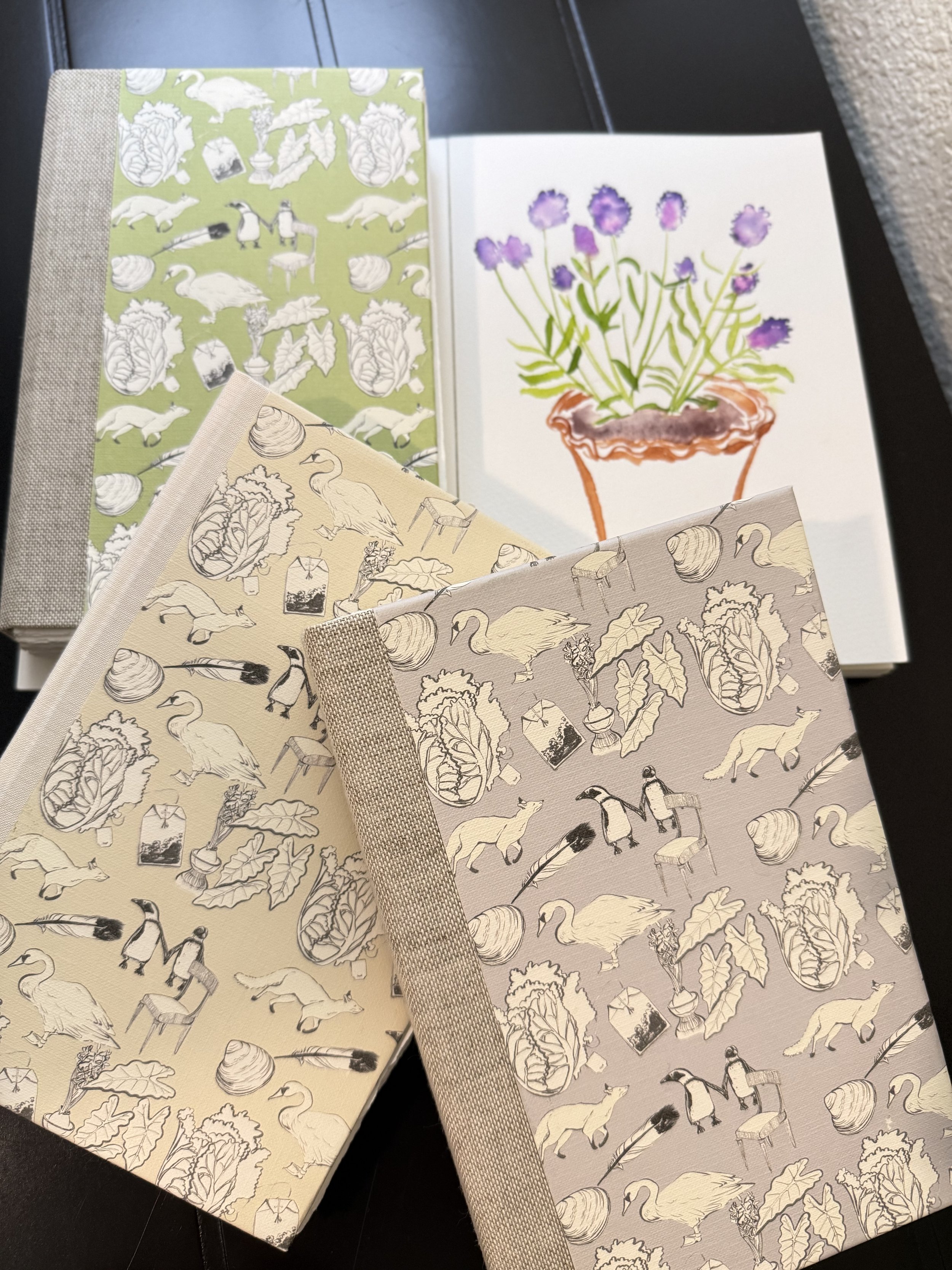 Three patterned notebooks or journals with nature and animal illustrations, one with a purple floral drawing in a pot, on a black surface.