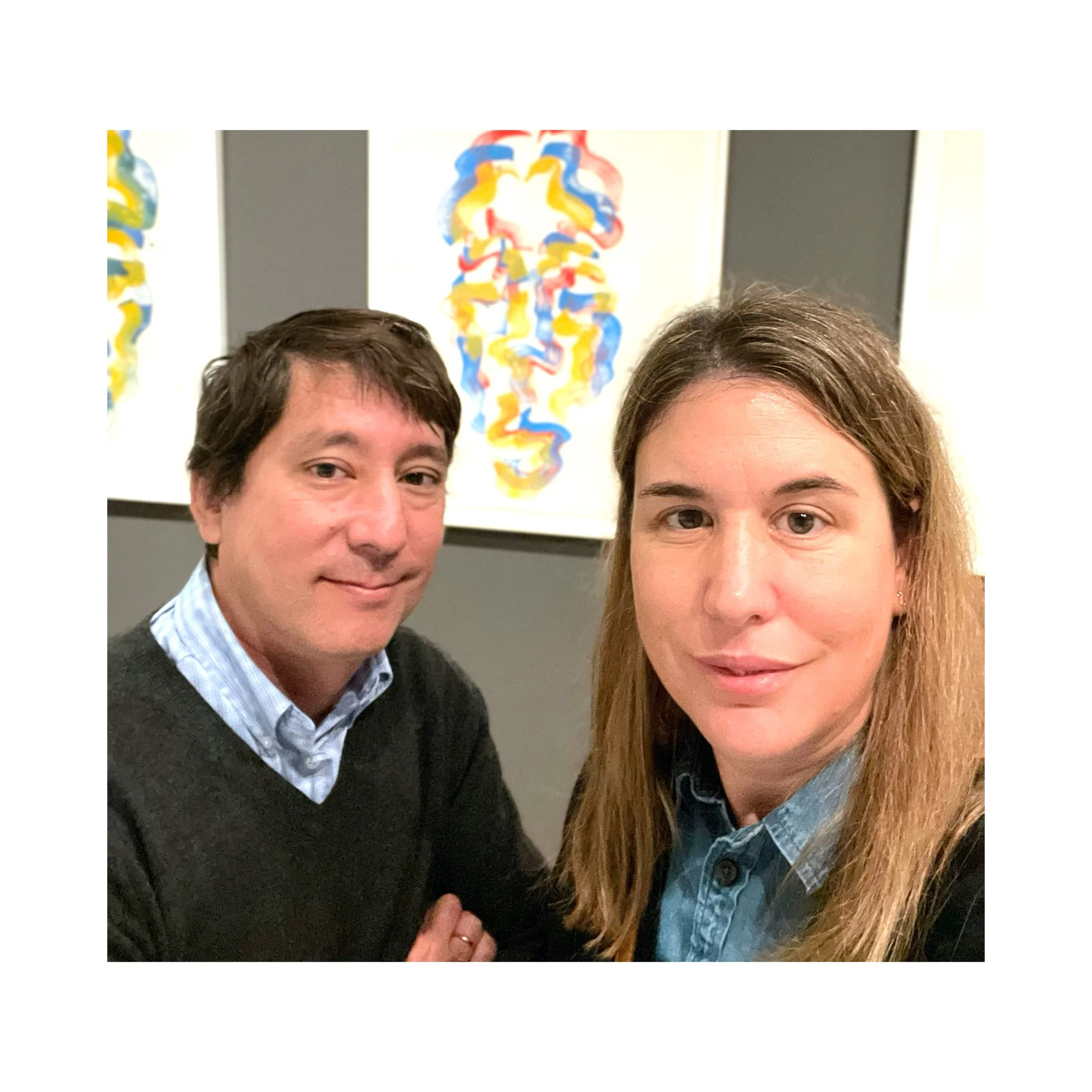 A man and woman take a selfie in front of colorful abstract artwork on a gray wall.