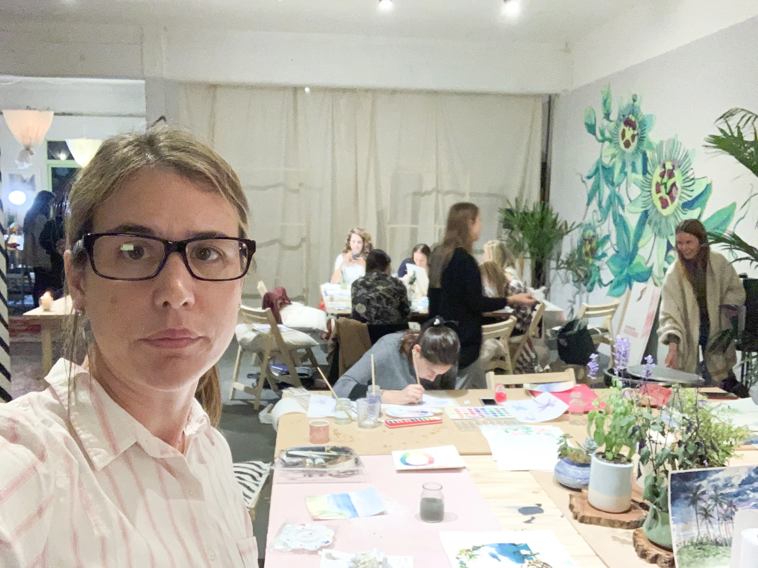 A woman wearing glasses and a striped shirt takes a selfie at an art workshop with people working on creative projects at tables in the background.