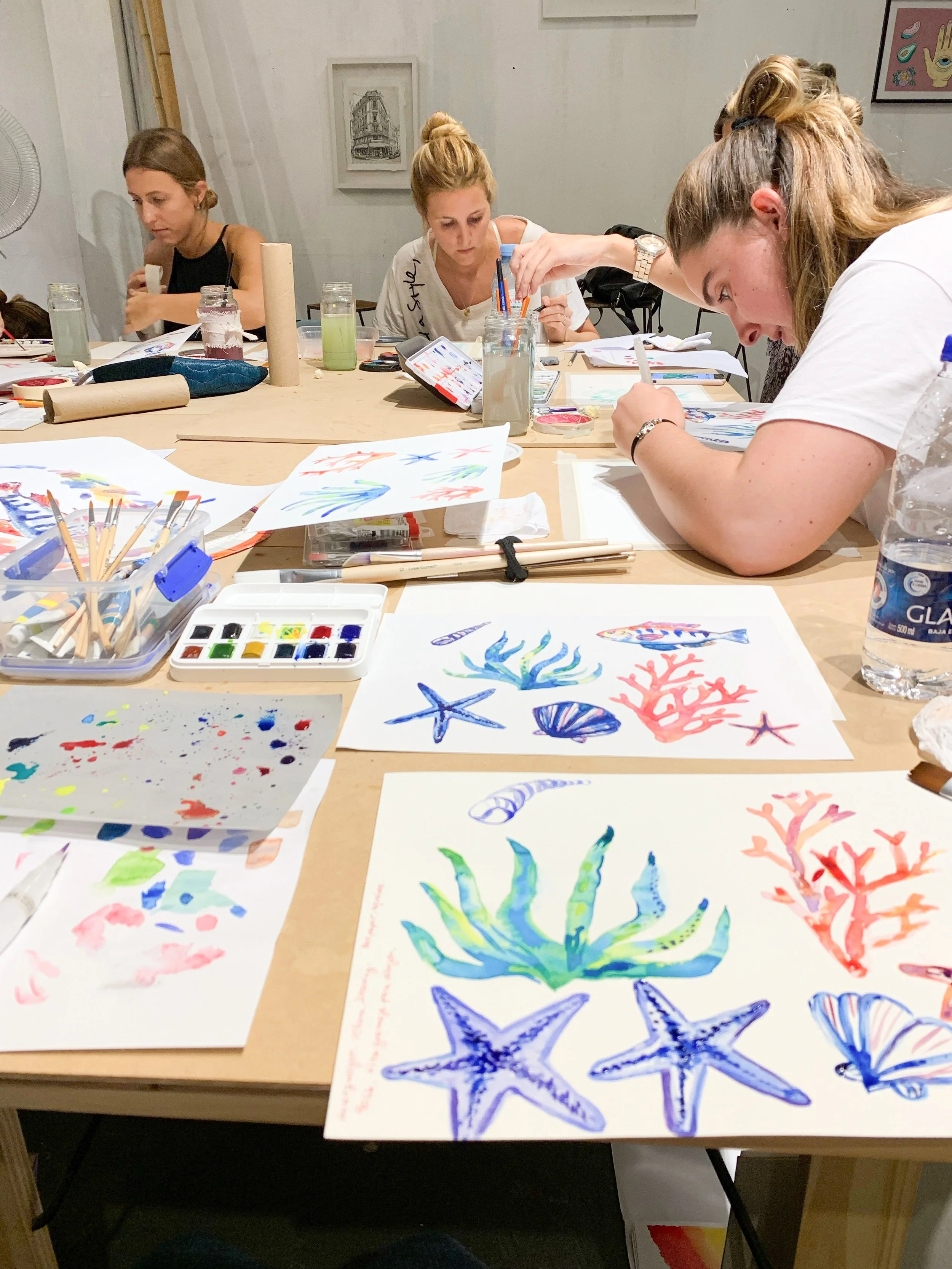 Women at a table painting colorful sea creatures like starfish, corals, and shells with watercolors during an art class.