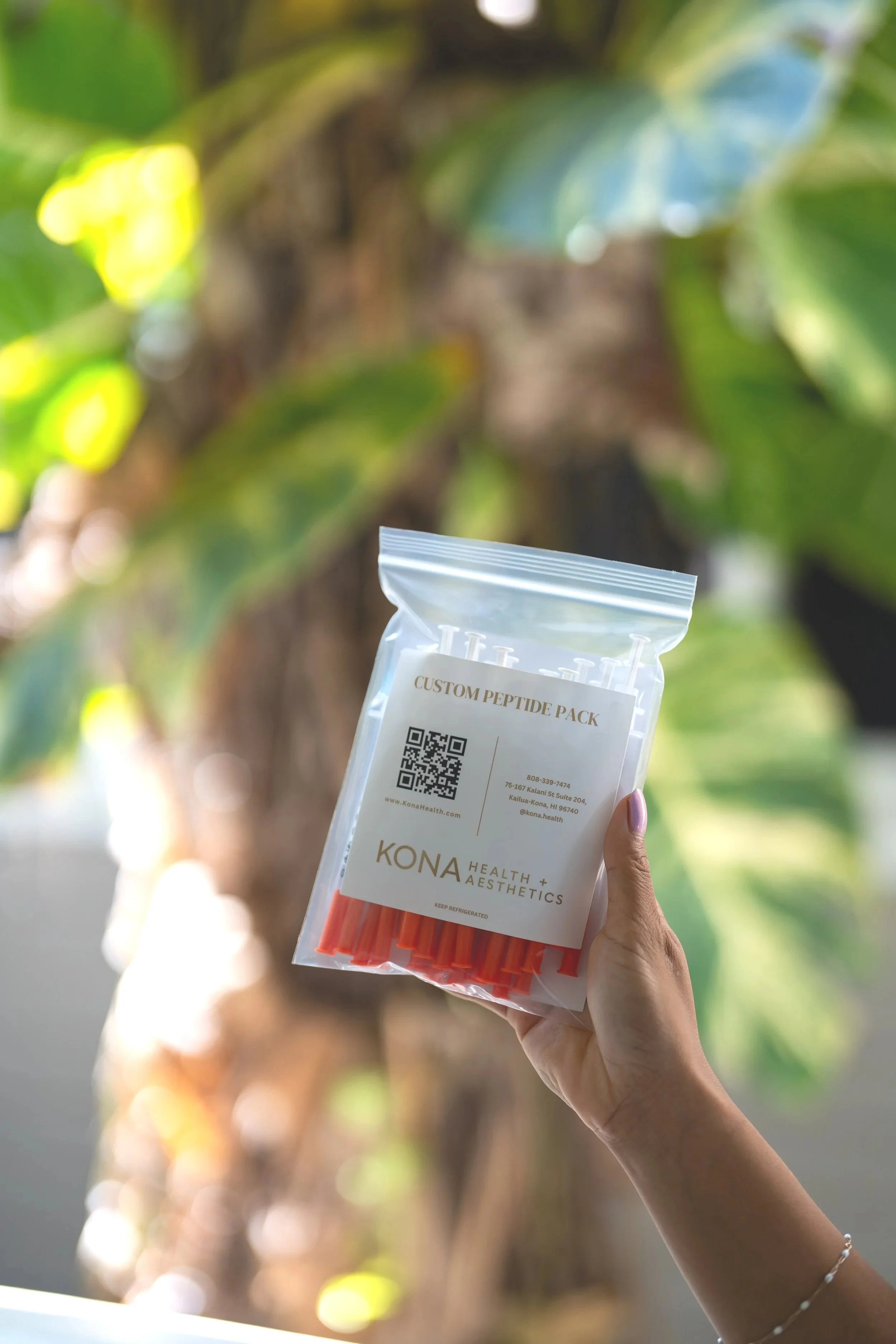 Hand holding up a Kona Health and Aesthetics Custom Peptide Pack against a soft blurred greenery background.