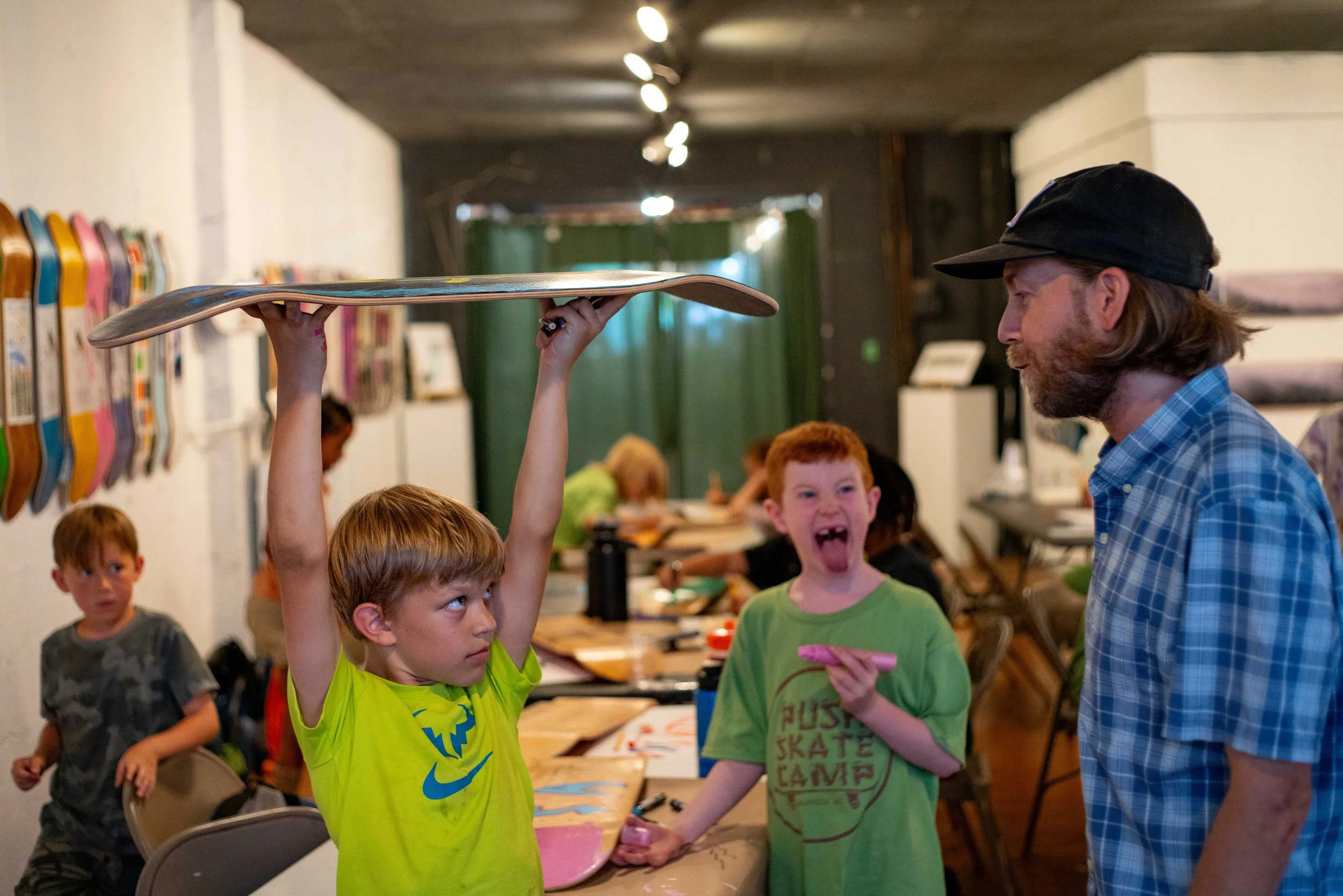 A young boy in a bright yellow shirt holds a skateboard overhead in a room full of kids and an adult male. The boy appears to be discussing or arguing, as another boy in a green shirt laughs and points at the skateboard. Several children are engaged 