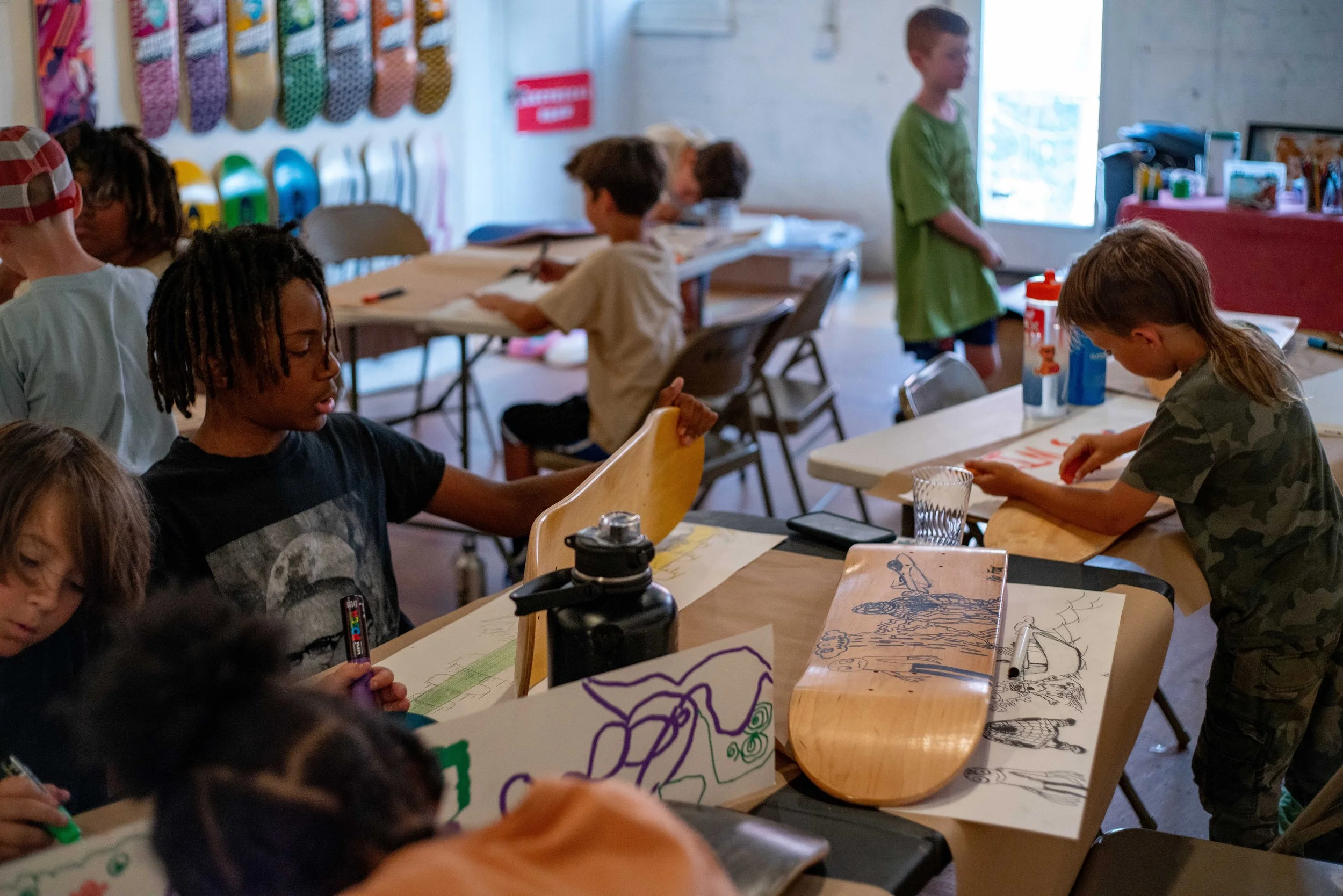 Children in a classroom engaging in art activities, drawing and coloring on paper and skateboard decks at tables.