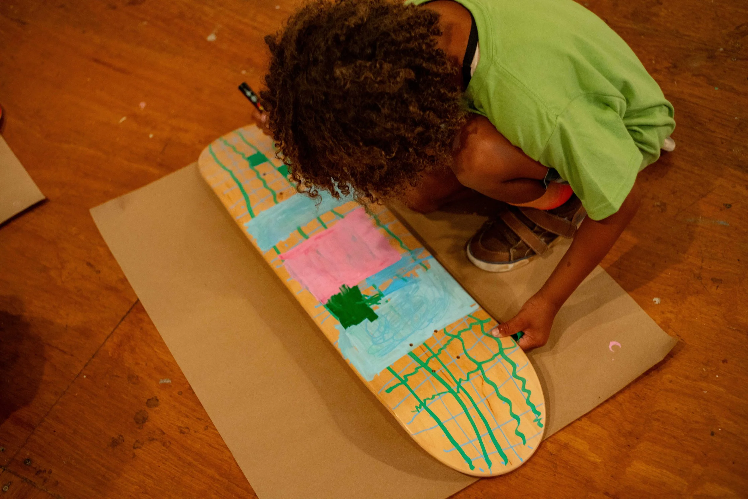 A boy with curly hair, dressed in a green shirt and brown shoes, is kneeling on a wooden floor, painting a colorful abstract design on a skateboard deck that is placed on a large piece of brown paper.