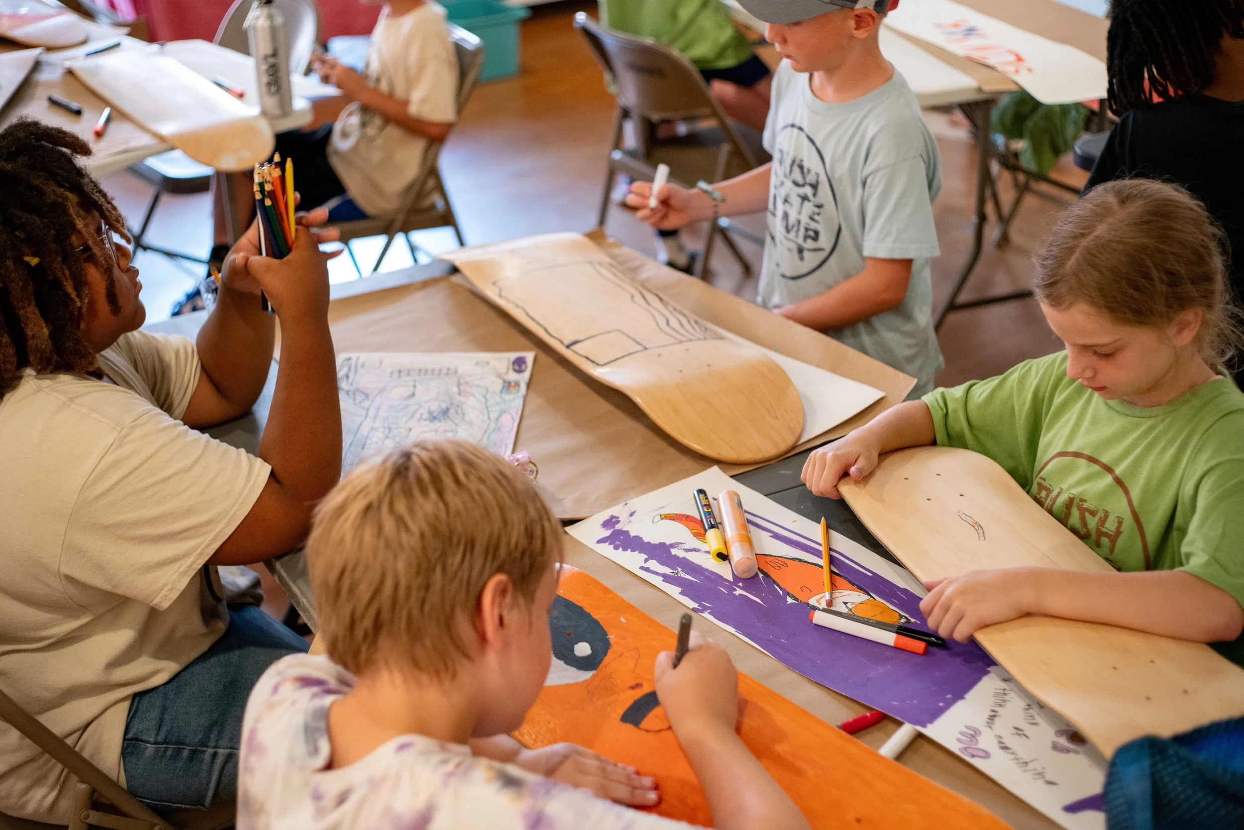 Children gathered around a table working on skateboard art projects, with markers, colored pencils, and artwork visible.