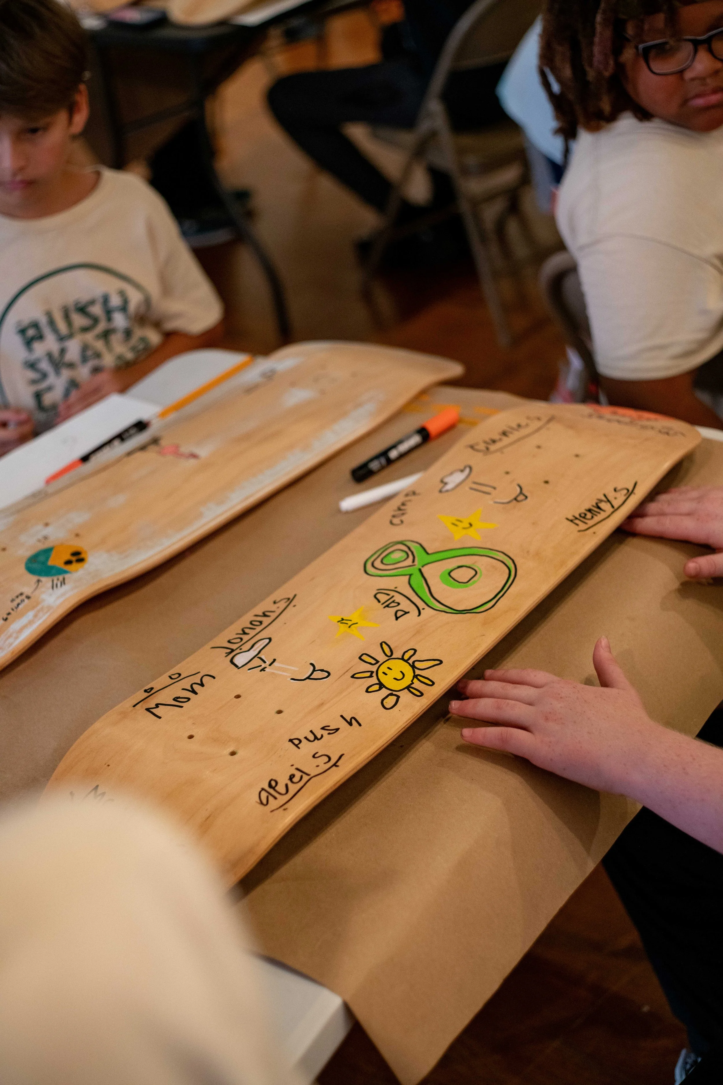 A decorated skateboard with colorful drawings and writings, including a smiling sun, a star, and a green spiral, placed on a table surrounded by children and adults during an arts and crafts activity.