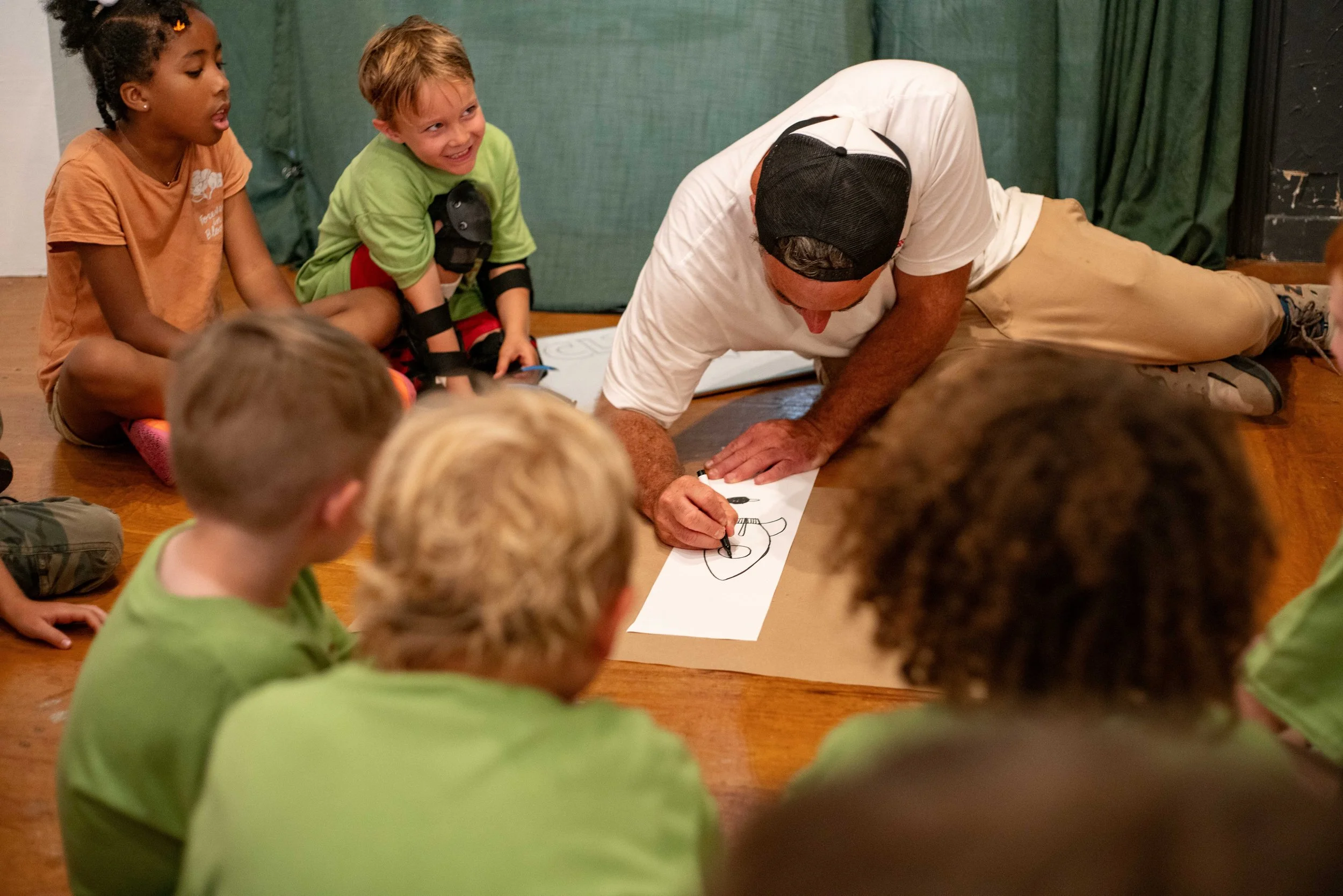 A man is drawing on a long piece of paper on the floor while children sit around watching. The children are smiling and appear engaged, some wearing green shirts. One child is dressed as a superhero with black armbands.