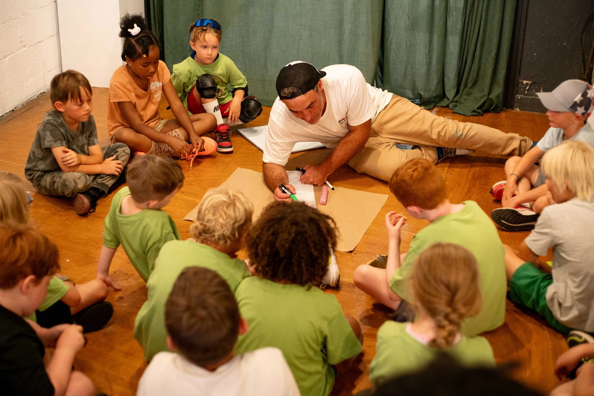 A man lying on the floor surrounded by children sitting in a circle on a wooden floor. The man is drawing on paper while the children watch.