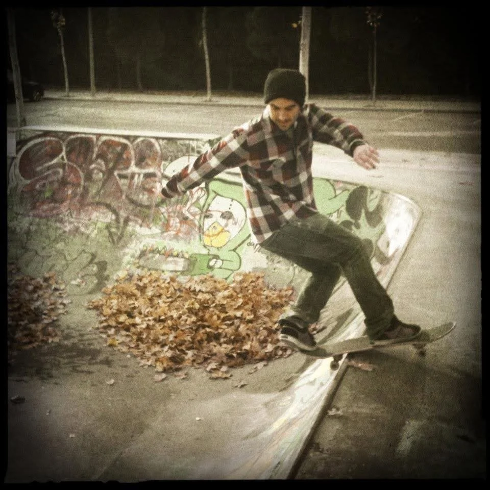 A young man wearing a beanie and plaid shirt skateboard tricks in a skate park at night, with leaves on the ground near a graffiti-covered ramp.