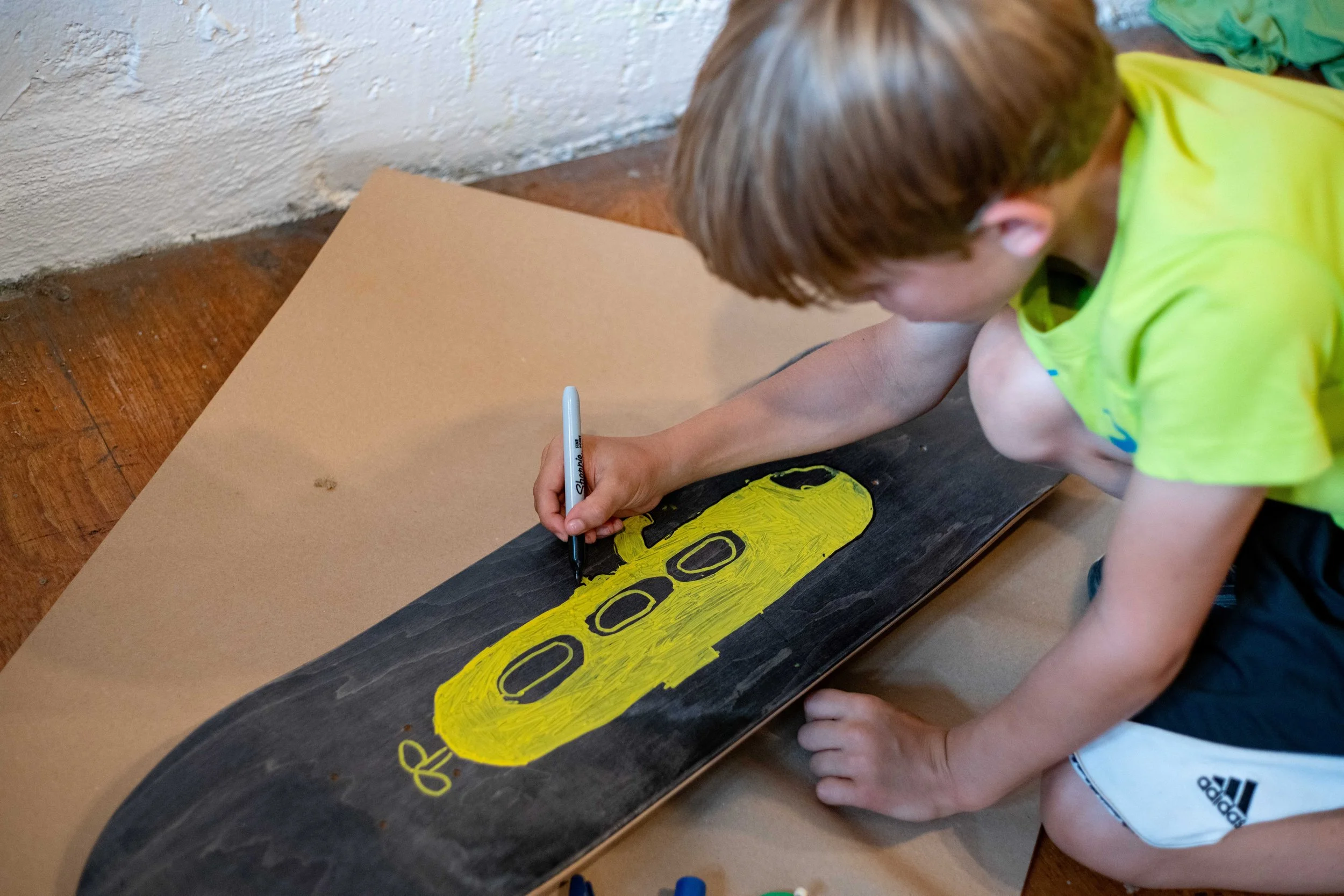A young boy in a green shirt and black shorts is drawing a yellow shape with black details on a skateboard with a black surface.