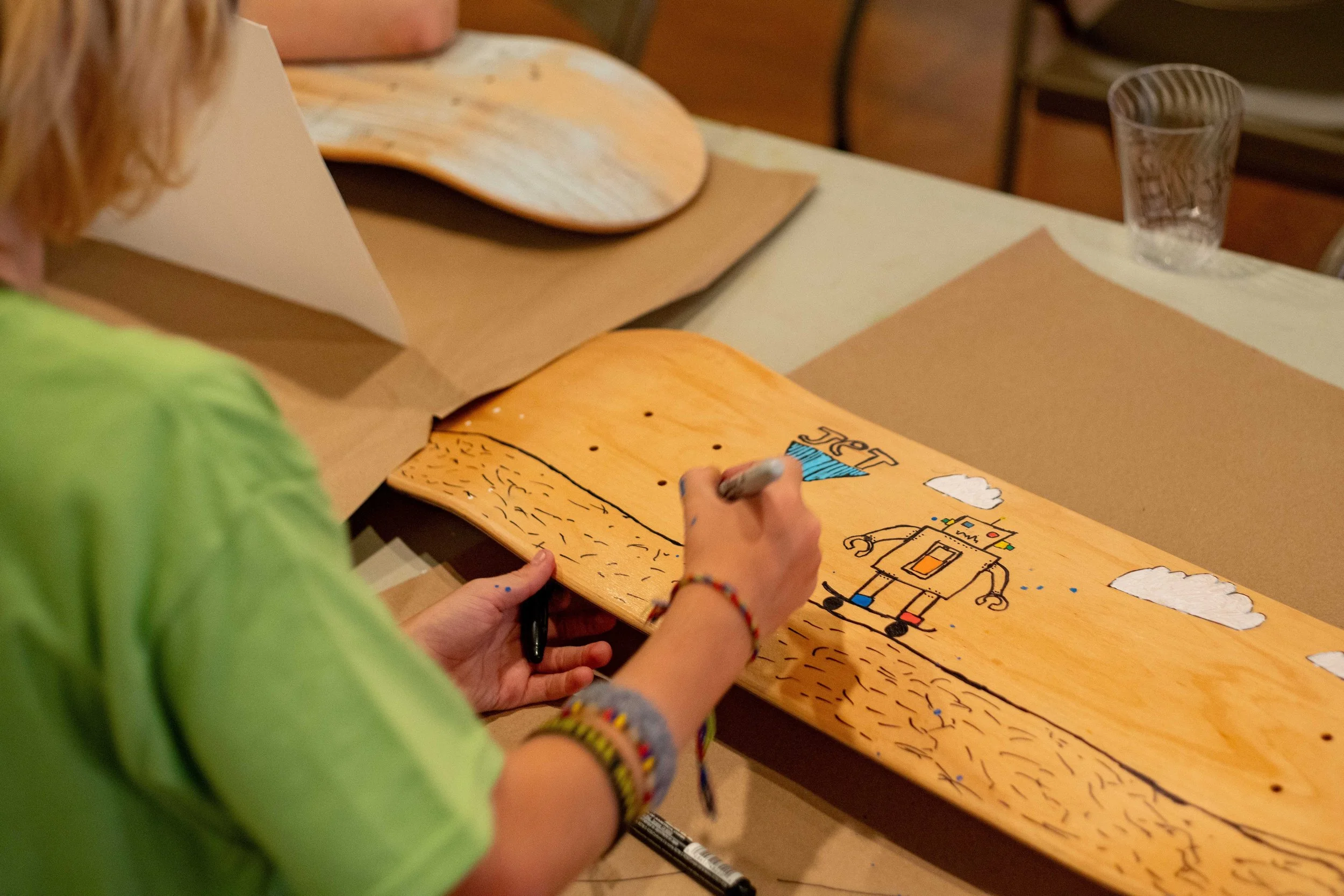 A person wearing a green shirt and colorful bracelets is drawing on a piece of wood with a black marker. The drawing depicts a robot, clouds, and a landscape.